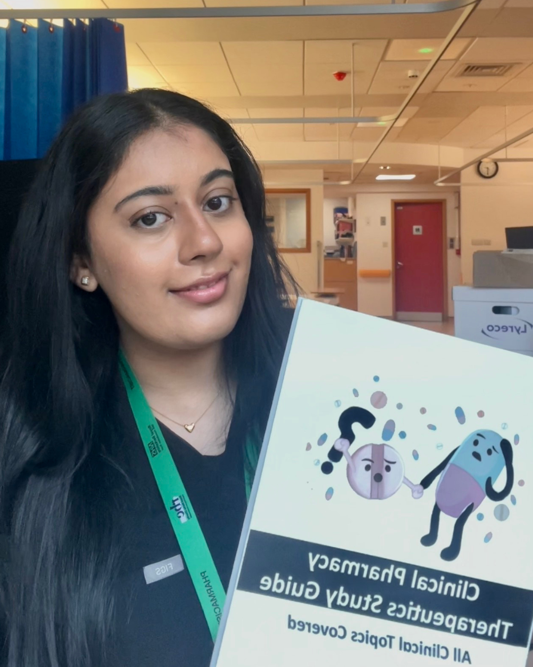 A woman with black hair, wearing earrings and a black top, holding a clinical pharmacy therapeutics study guide, smiling at the camera in a hospital or clinical setting.