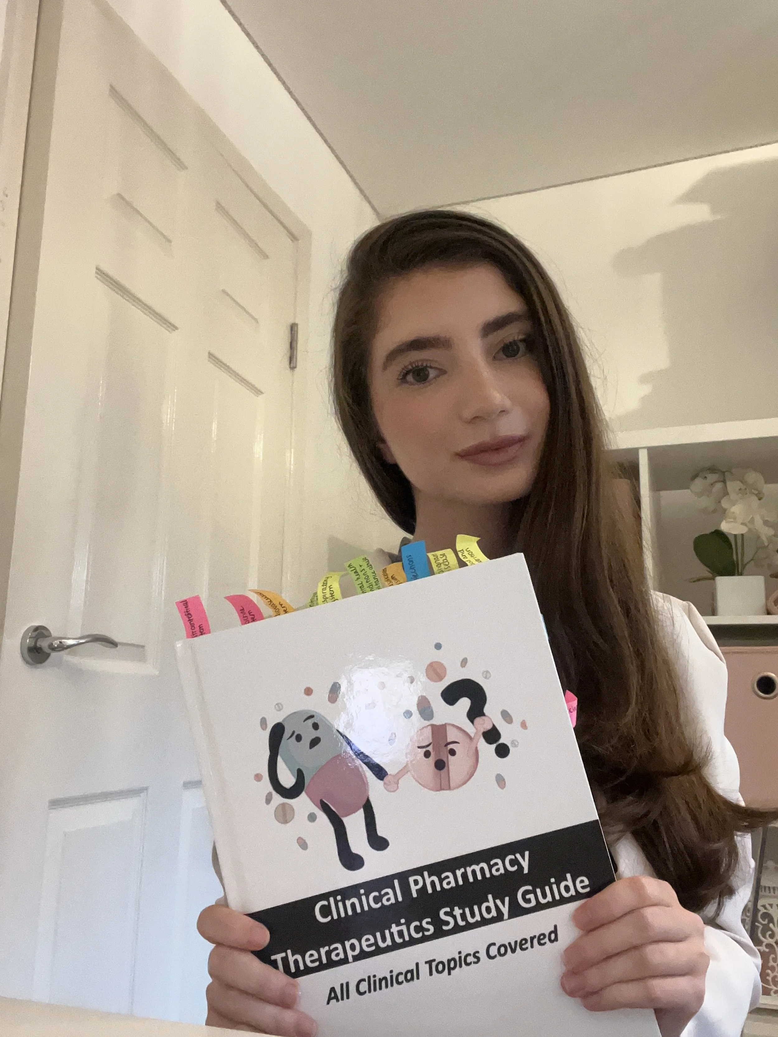 A young woman with long brown hair holding a clinical pharmacy therapeutics study guide with colorful tabs, standing in a room with white walls and a door.