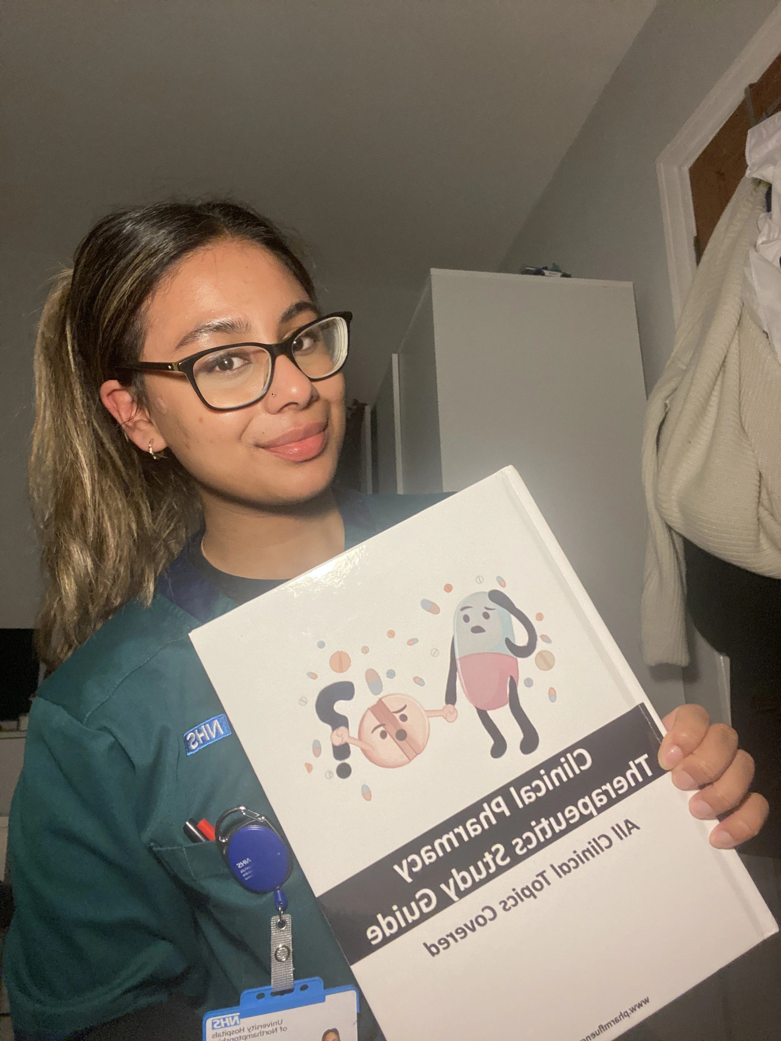 A nurse holding a booklet titled 'Clinical Pharmacy Therapeutics Study Guide.' The nurse is smiling and wearing a green uniform with an ID badge and name tag.