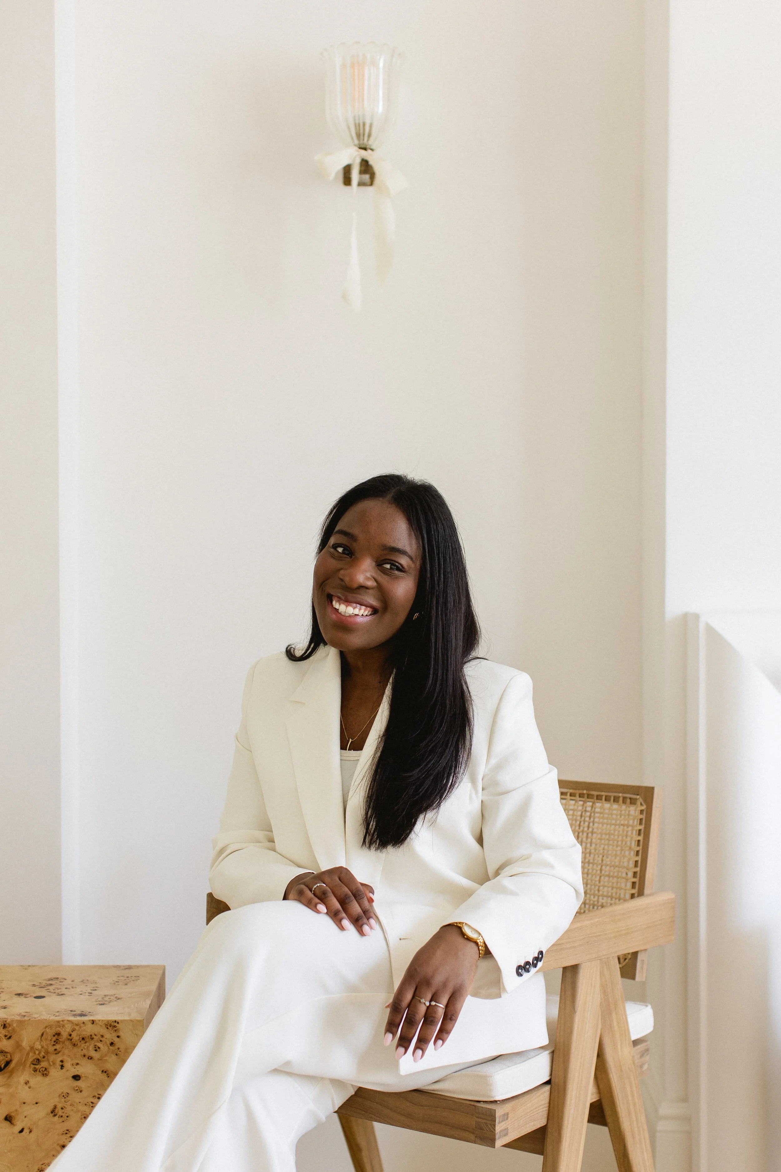 A woman in all-white attire sitting on a wooden chair smiling at the camera, with a light-colored wall and a wall-mounted light fixture behind her.