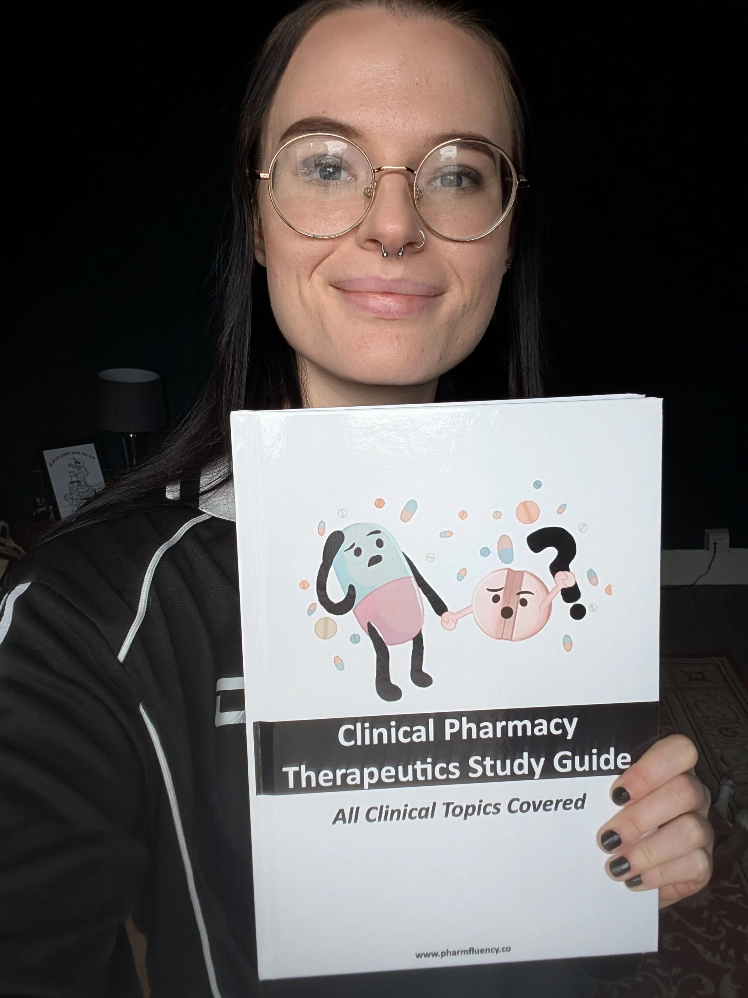 A woman with glasses and a septum piercing smiling, holding a book titled "Clinical Pharmacy Therapeutics Study Guide" with cartoon characters on the cover.