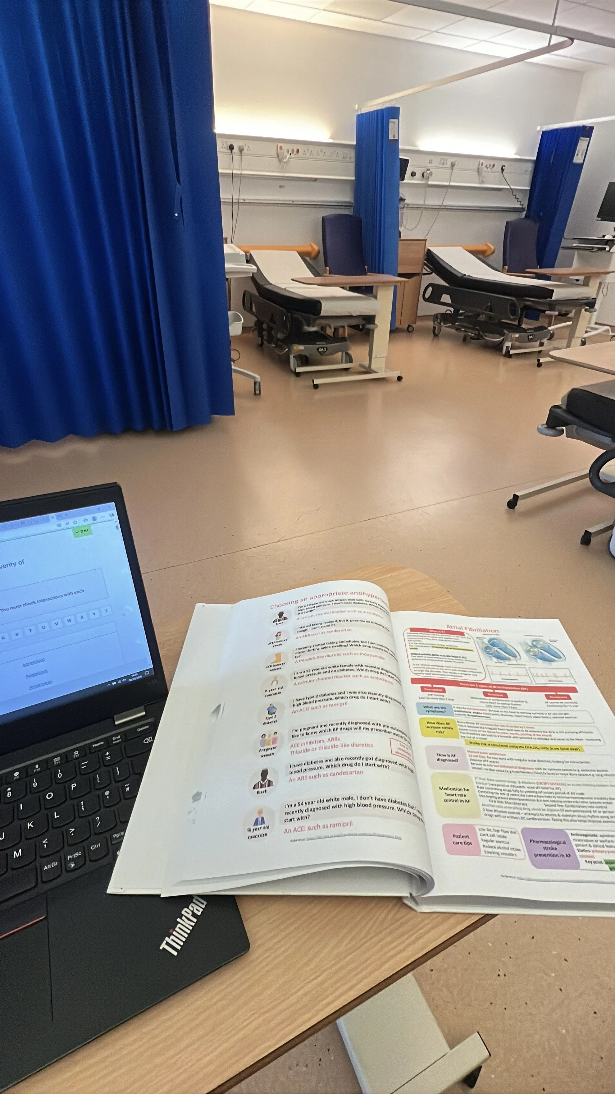 Hospital room with multiple beds separated by blue curtains, medical equipment along the wall, and a desk with an open medical textbook and a laptop.