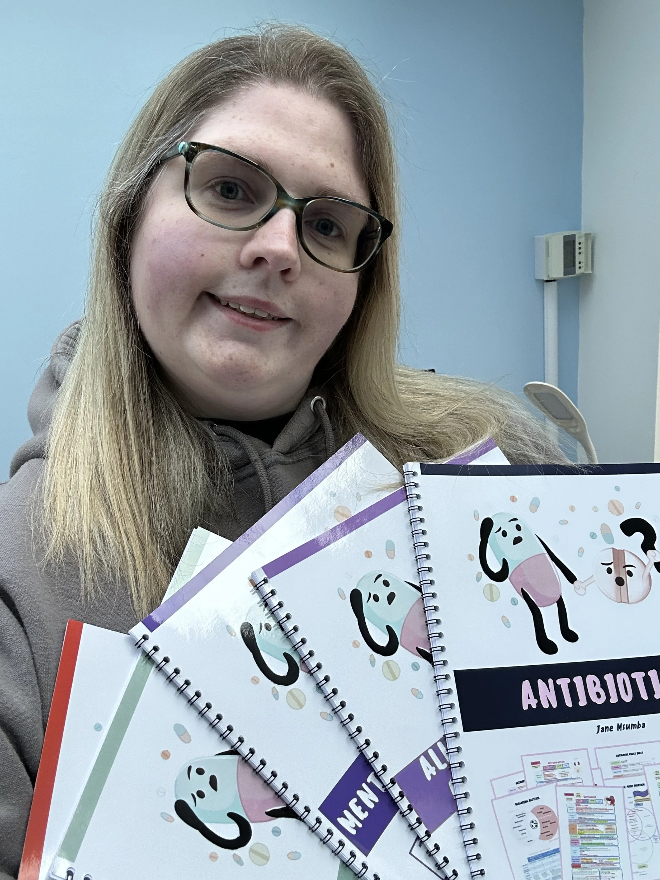 Woman with glasses holding multiple notebooks titled 'ANTIBIOTICS' with cartoon characters and diagrams, in a room with blue walls and a thermostat in the background.