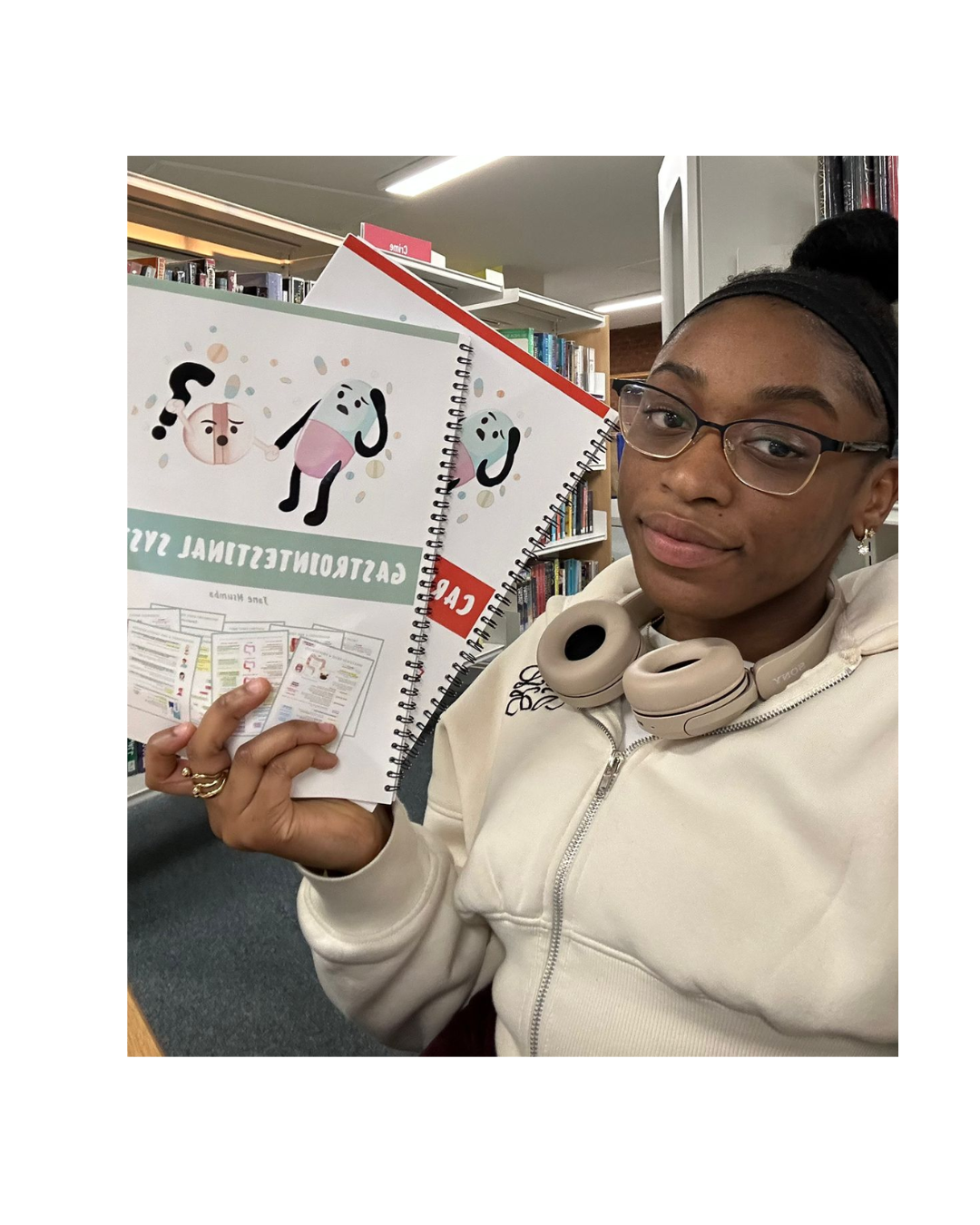 A young woman with glasses and a beige hoodie holding books and notebooks in a library.