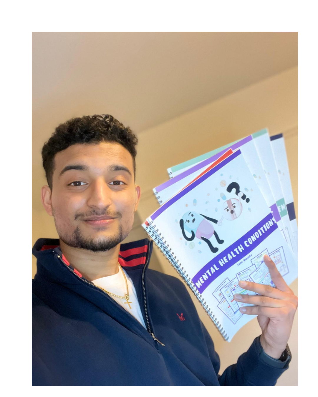 A young man with short curly hair and a beard holding a notebook titled 'Mental Health Conditions' with cartoon illustrations of mental health characters, smiling at the camera, indoors against a beige background.
