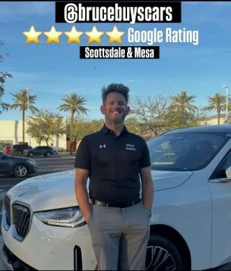 A man standing next to a white car with a clear blue sky and palm trees in the background. He is smiling and wearing a black polo shirt and gray pants. The image has a graphic overlay with social media handle '@brucebuyscars,' a five-star Google rating, and mentions Scottsdale & Mesa.