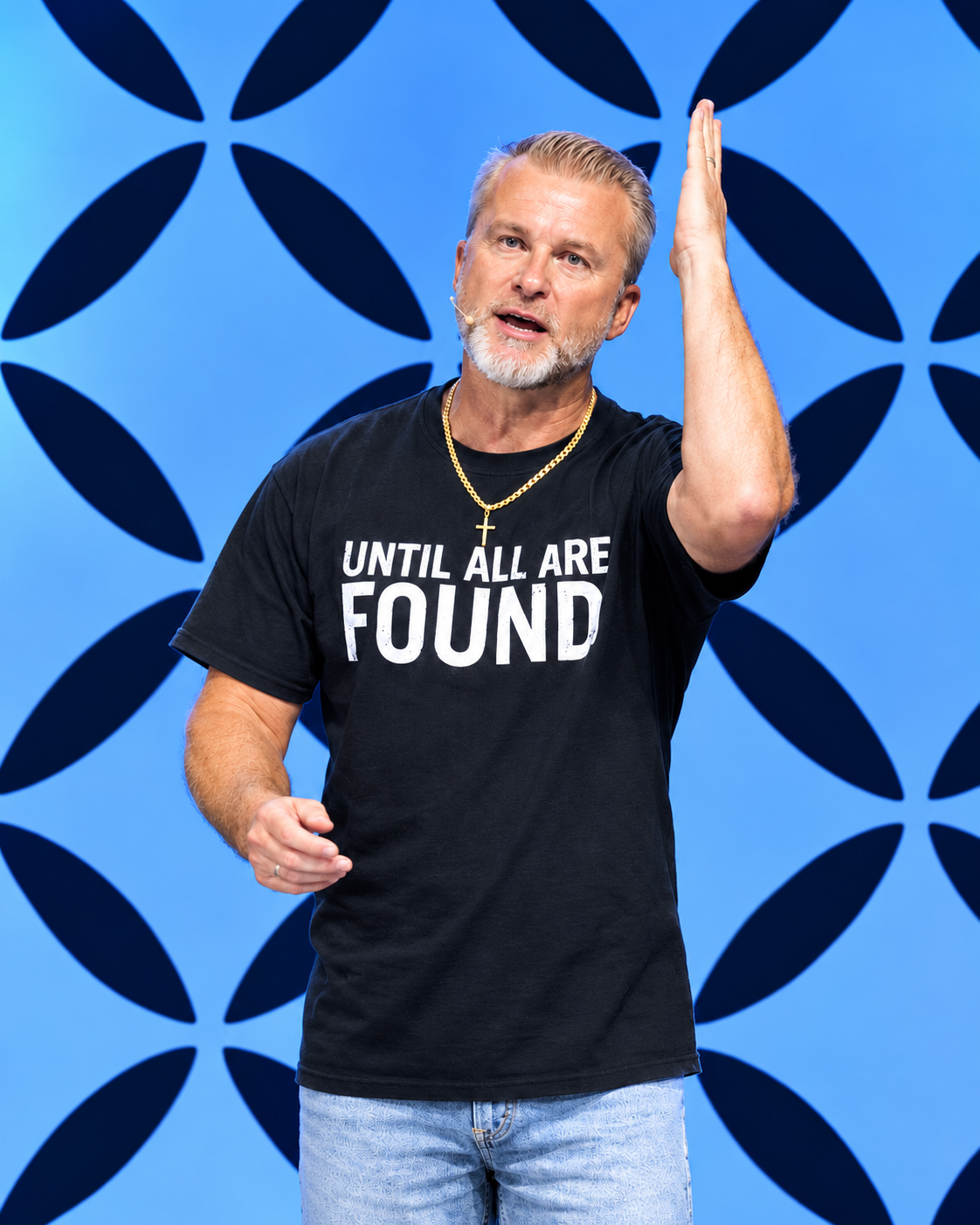 A man with gray hair and a beard speaking on stage, wearing a black t-shirt that says 'Until All Are Found' and a gold cross necklace, with a blue geometric patterned background.