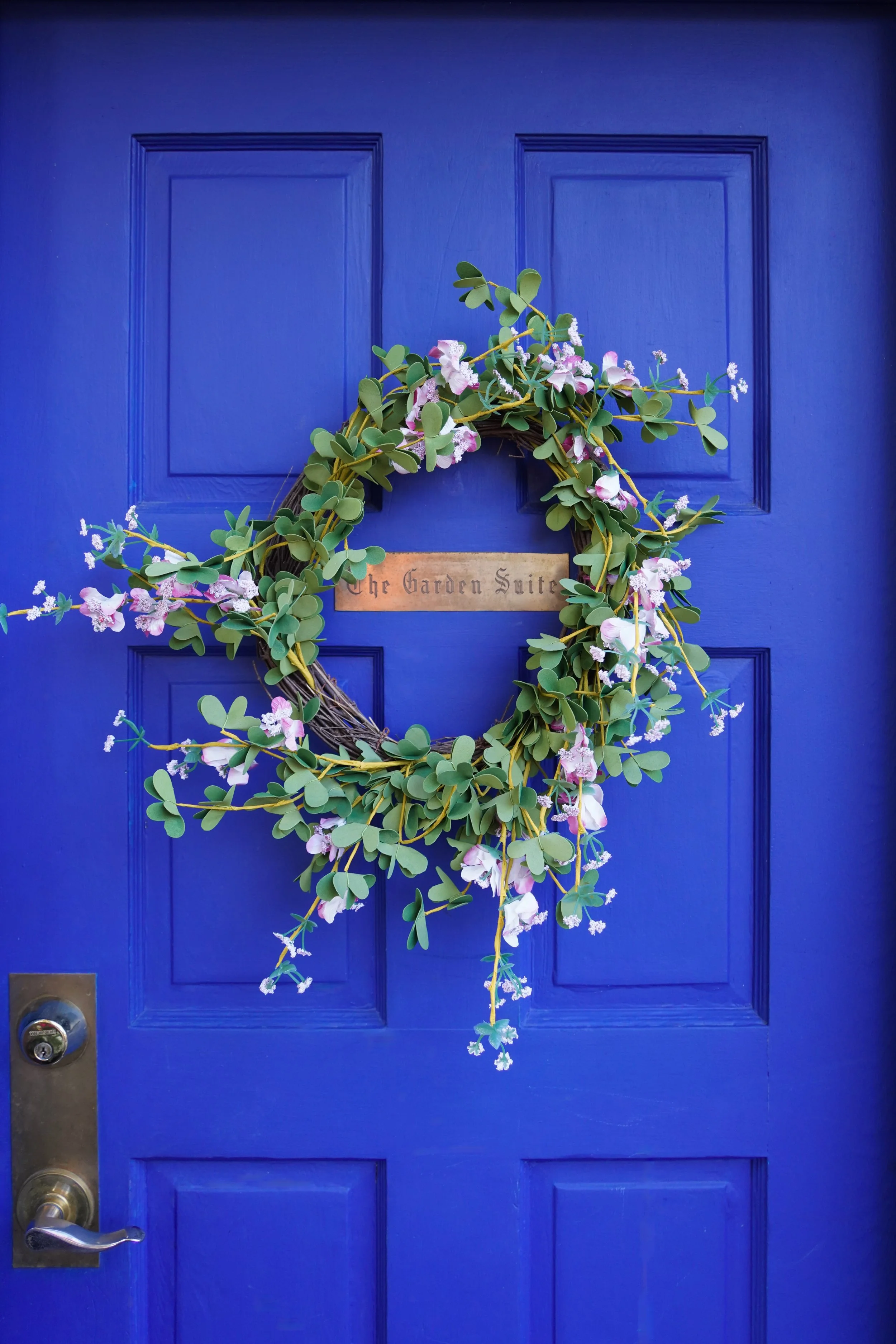 A blue door with a decorative wreath made of green leaves and pink and white flowers, with a wooden sign that reads 'The Garden Suite' in the center.