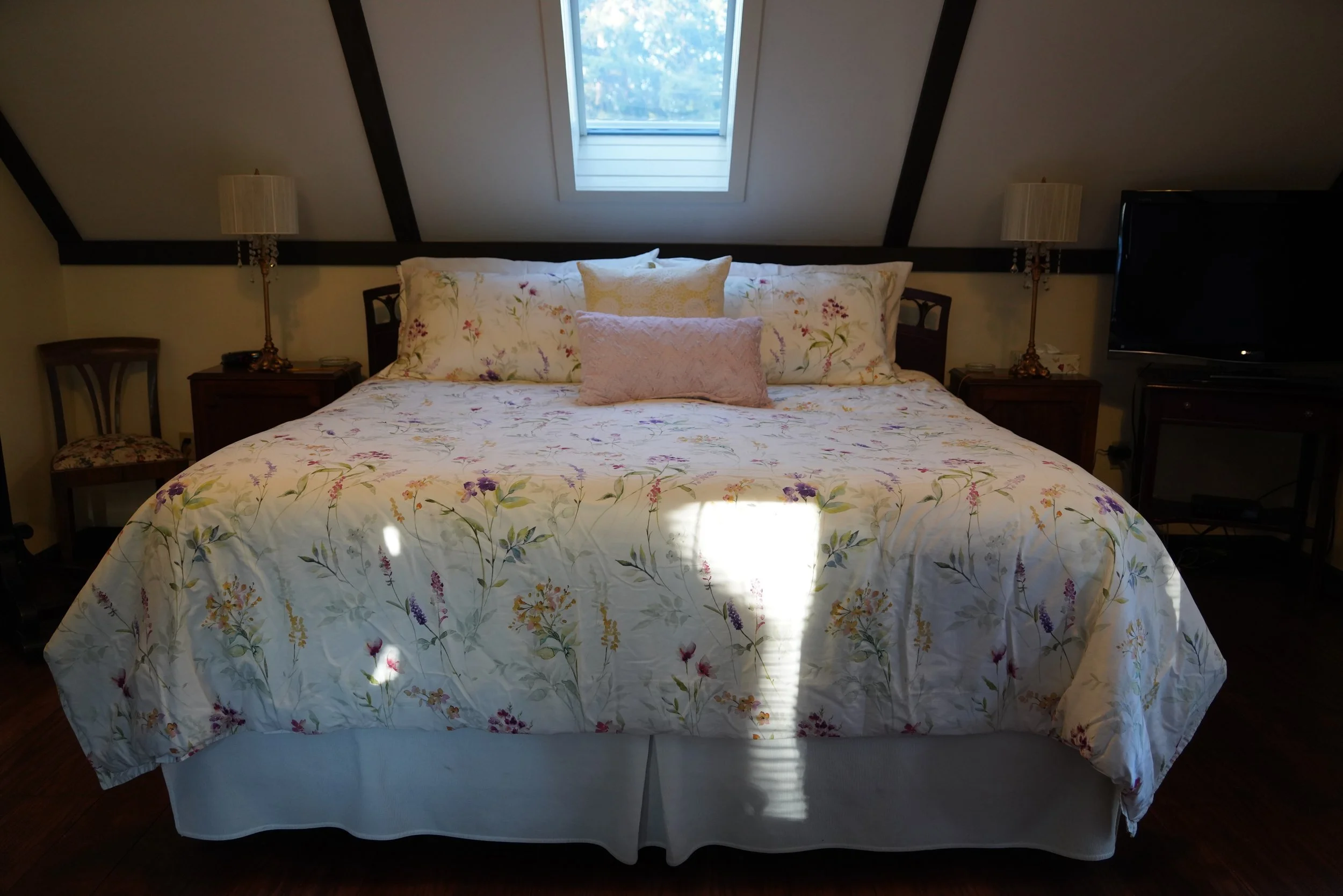 A bedroom with a bed featuring a floral bedspread, two pillows, and a nightstand on each side with lamps. There is a window in the slanted ceiling above the bed.