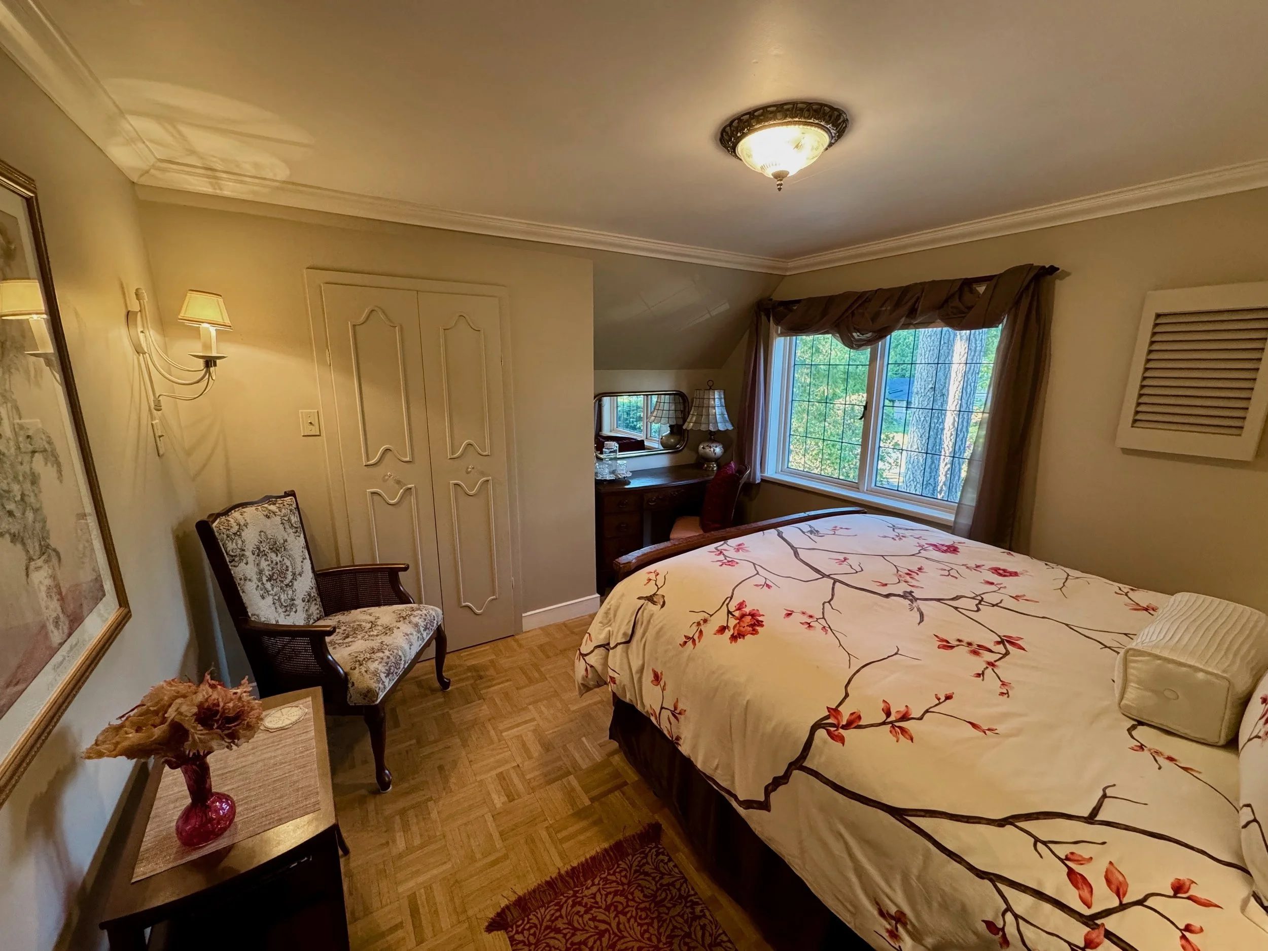 A cozy bedroom with a large bed covered in a floral-patterned duvet, a window with curtains, a wooden desk with a mirror and lamps, a chair with a floral cushion, a small table with a vase of flowers, and a ceiling light fixture.