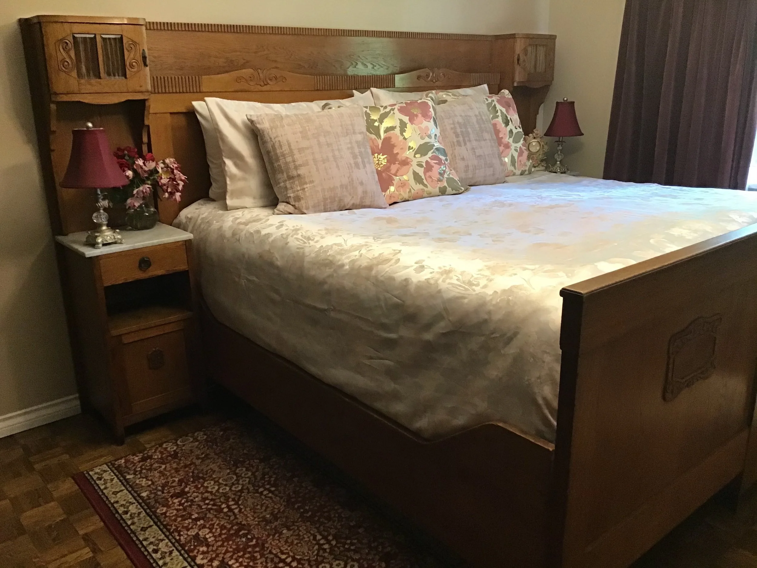 A neatly made bed with a wooden headboard and footboard in a bedroom. The bed has beige, floral, and plaid pillows, and a floral-patterned bedspread. There are two nightstands with red lamps and decorative items, and a small rug on a wooden floor in front of the bed.