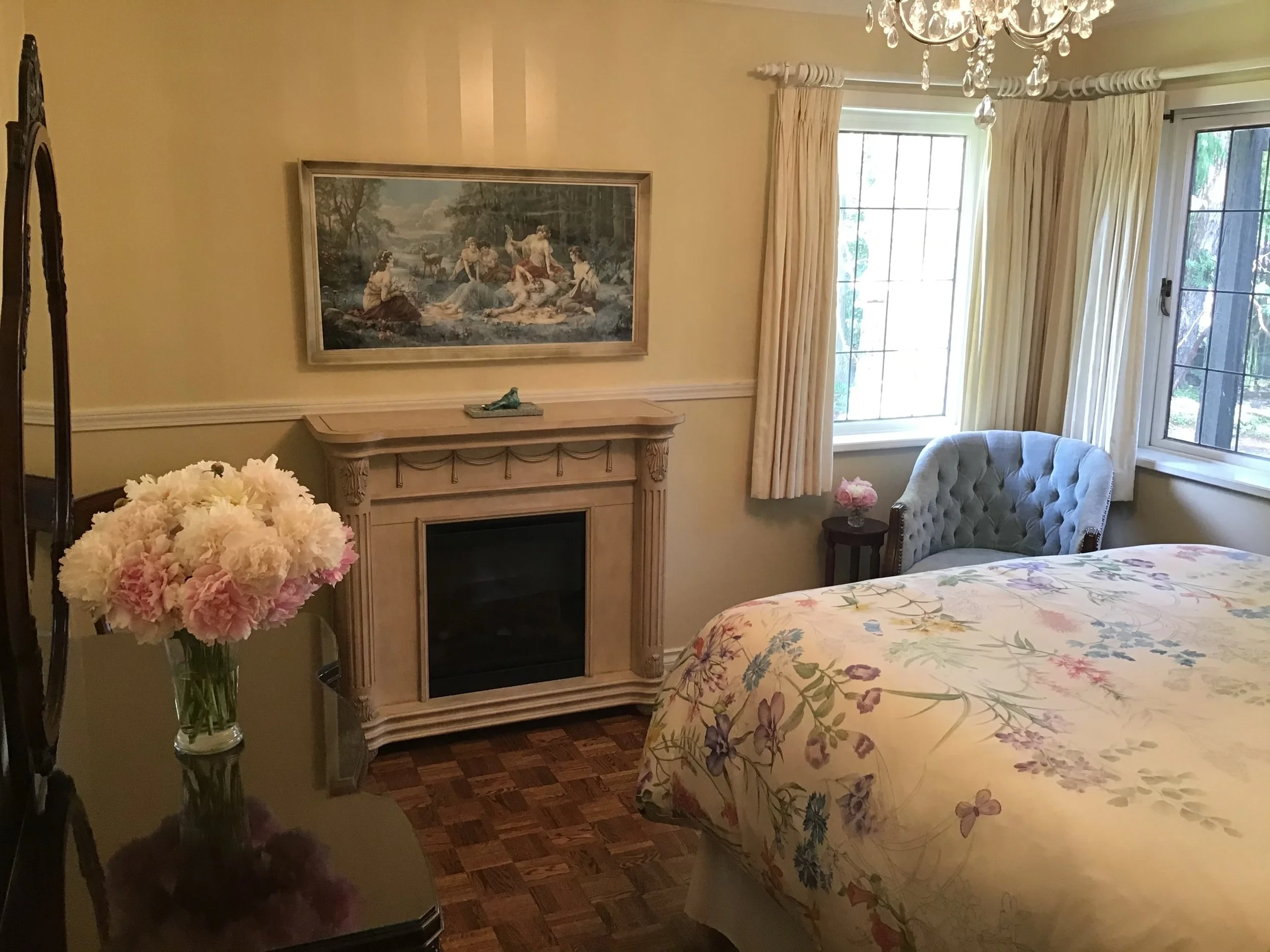 A cozy bedroom with a floral bedspread, a fireplace with a painting above it, a blue armchair near a window, and a small side table with a pink flower arrangement.