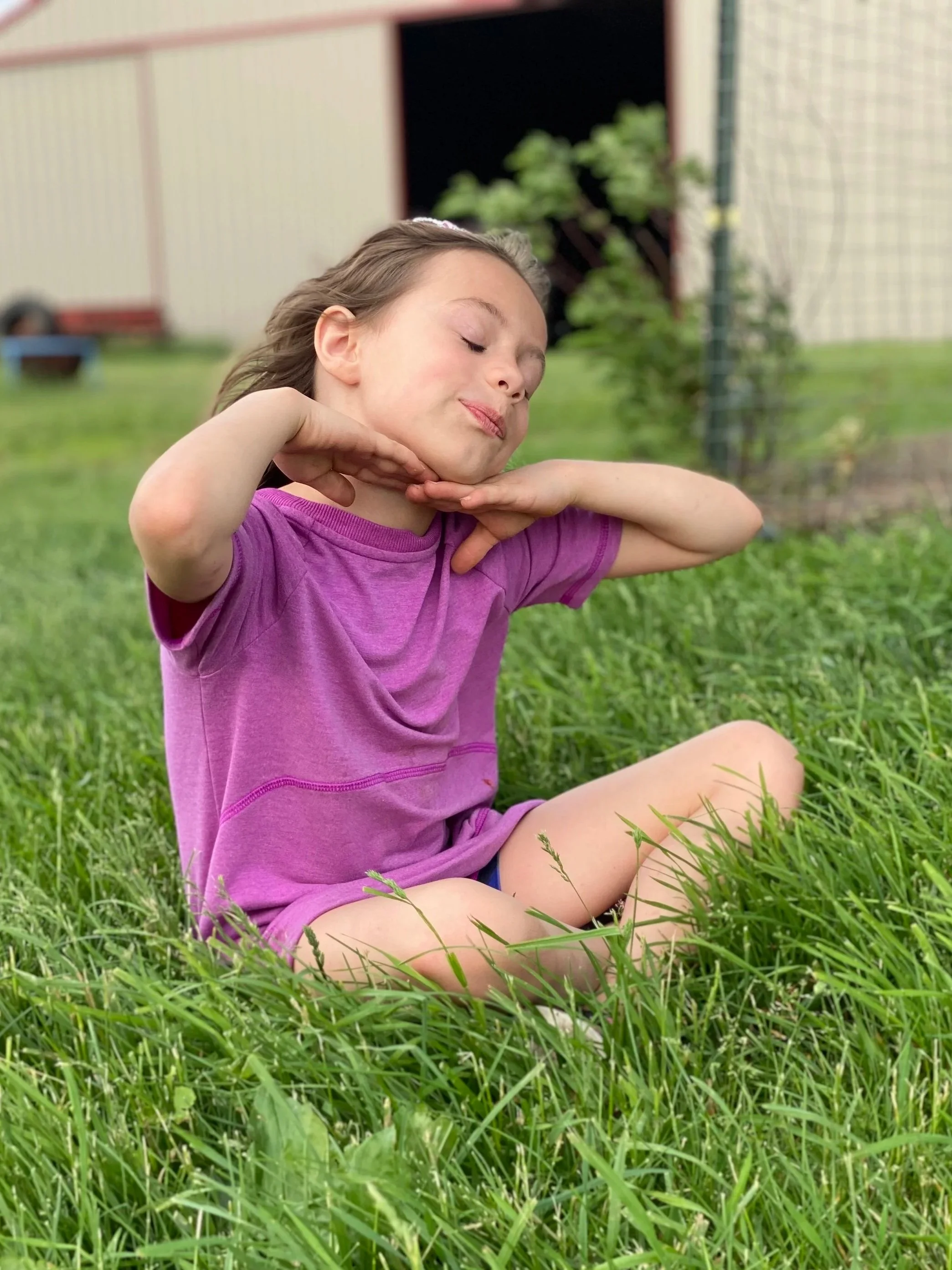 A young special needs girl sitting cross-legged in a grassy yard with her eyes closed, hands under her chin, and a content smile on her face, wearing a purple T-shirt.