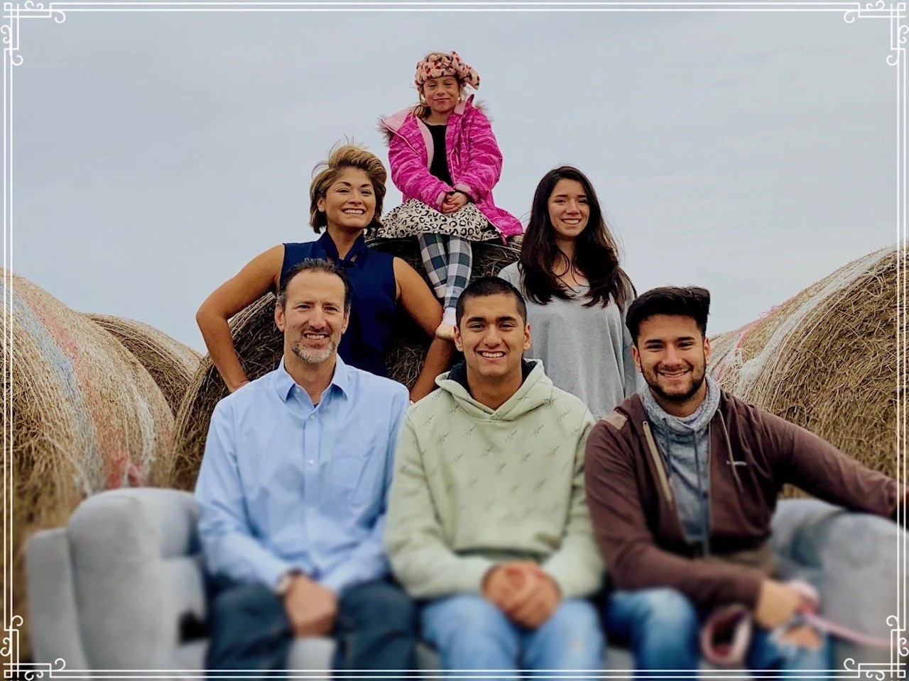 A family of six, including two women, two men, and two young girls, sitting and standing on a hay bale in a rural setting with a cloudy sky in the background.