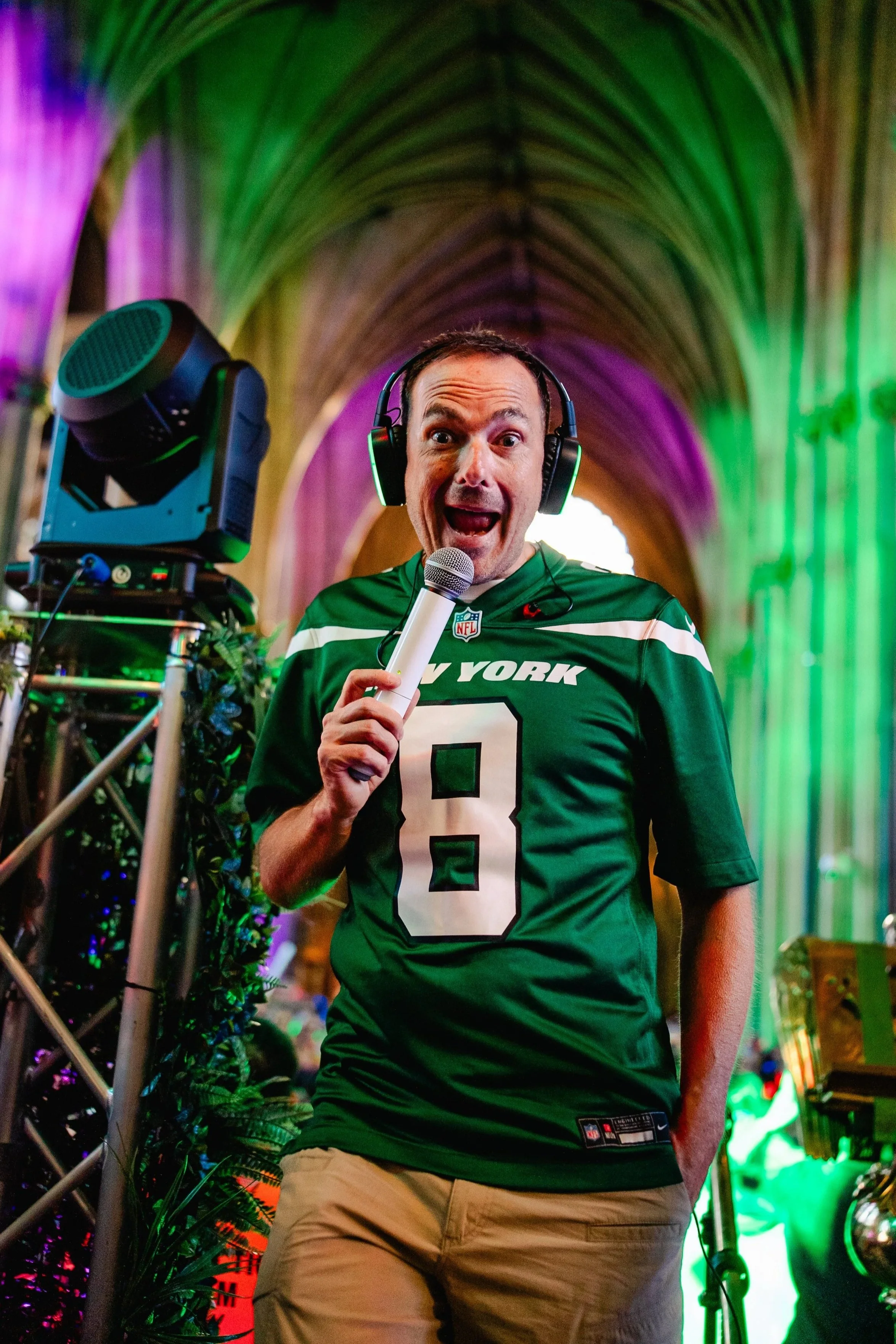 A man in a green New York football jersey, wearing headphones, holding a microphone, speaking excitedly with a surprised expression in a colorful, arched, possibly cathedral-like setting.