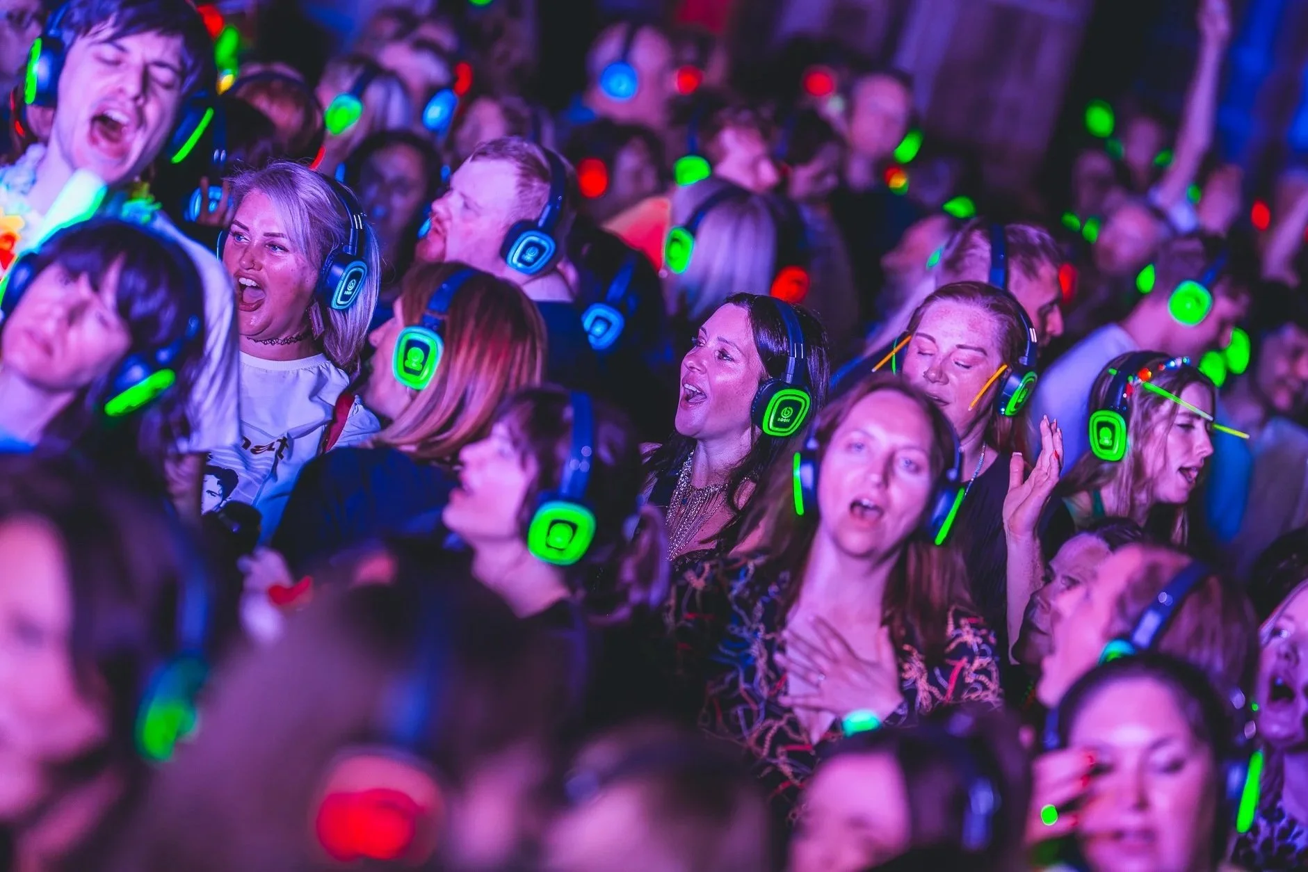 Crowd of people wearing neon headphones and singing along at a concert or dance event, with colorful lighting.