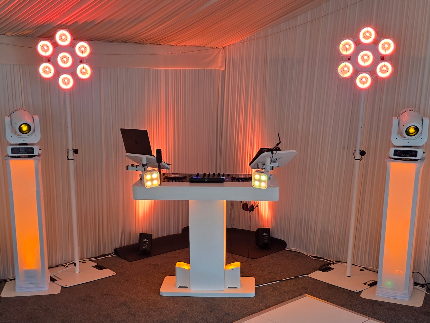 A DJ setup with two turntables on a white stand, a laptop, and various lighting equipment including two large hexagon-shaped lights and two tall stand-mounted lights with yellow and orange accents, set against a draped wall with warm orange lighting.