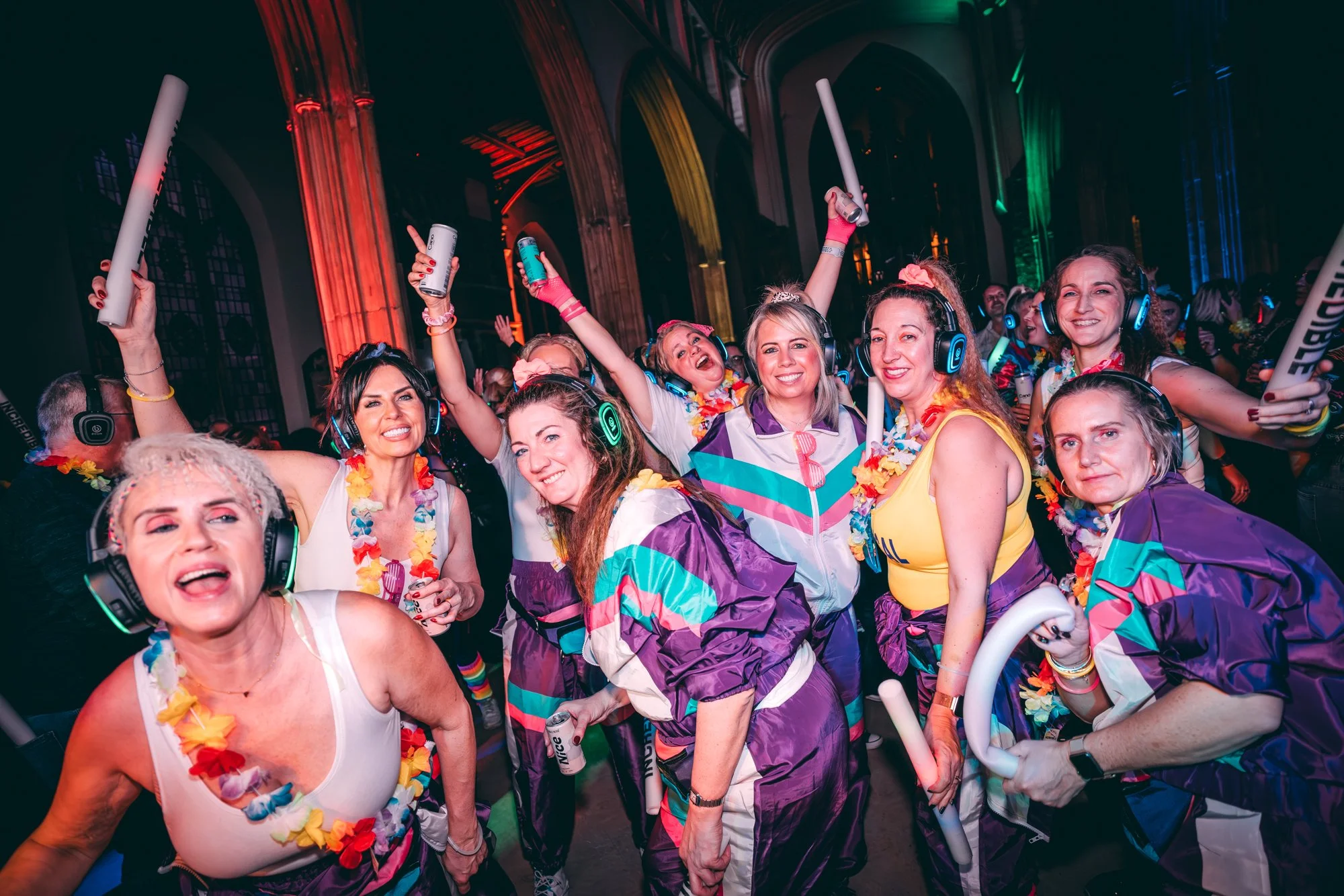 Group of women at a lively indoor party wearing colorful 80s-style outfits, headphones, leis, and holding glow sticks, dancing and smiling with vibrant multicolored lighting in the background.