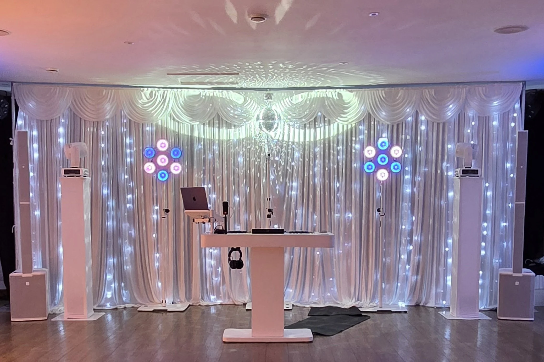 DJ booth setup with speakers, lighting, and a white backdrop with curtains and string lights, in a decorated event space.