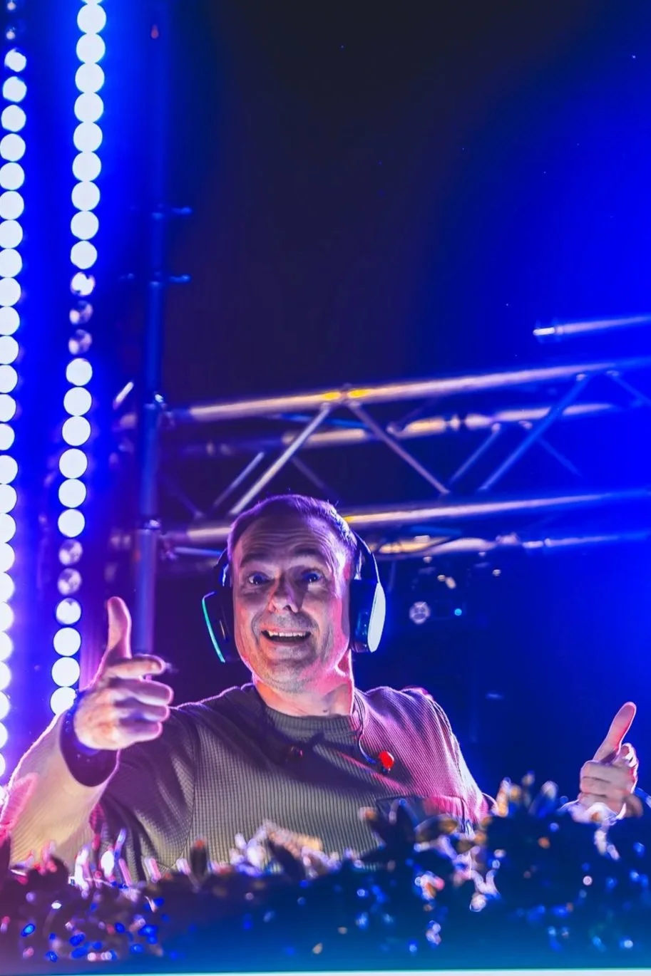 A DJ wearing headphones, smiling, and giving a thumbs-up at a music event, with colorful lighting and stage equipment in the background.