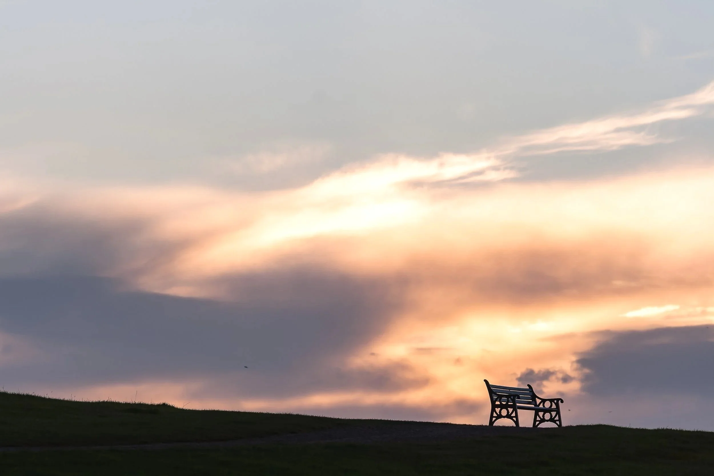 A wooden park bench on a grassy hill at sunset, with clouds in the sky.