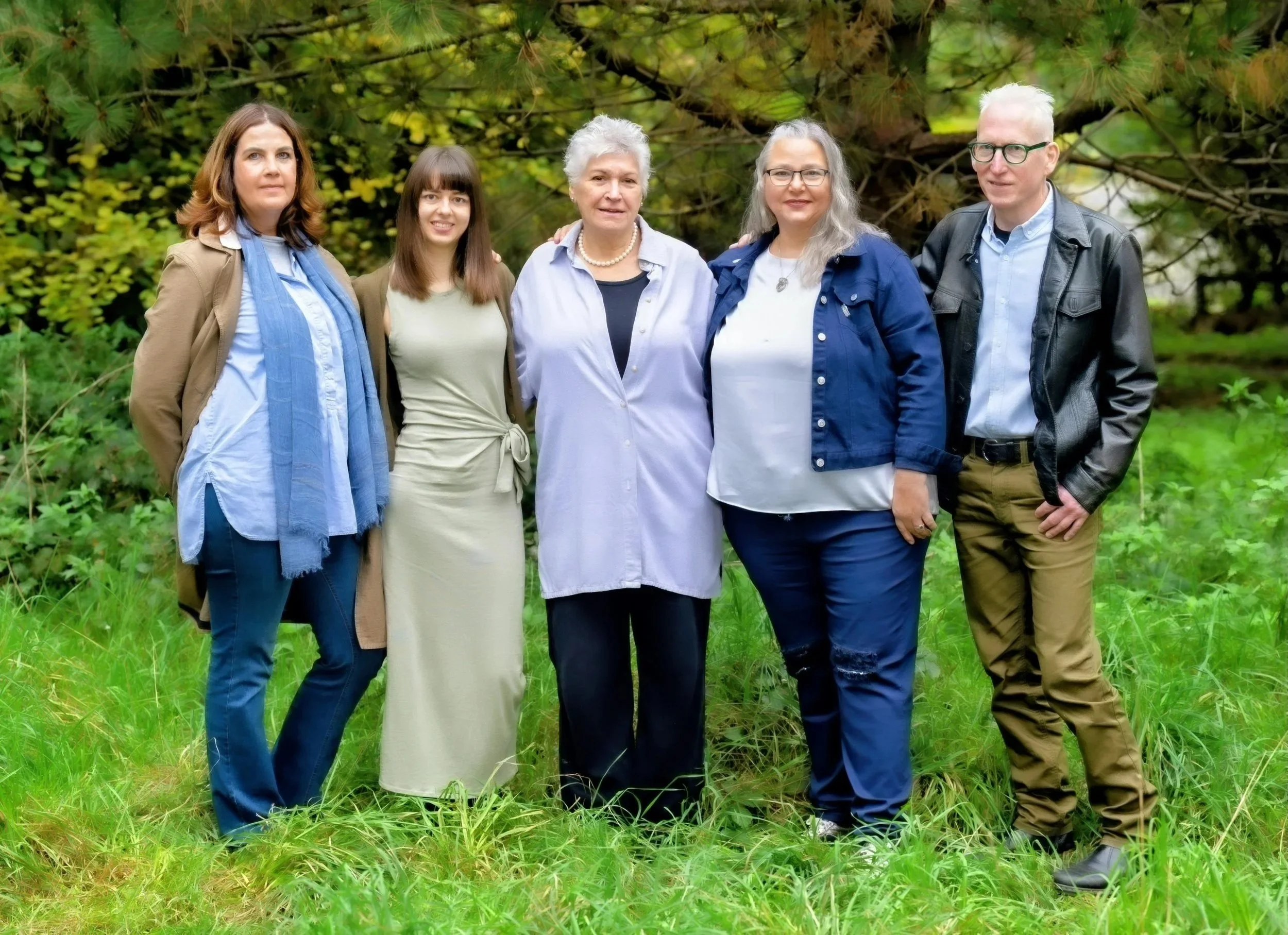 Group of five women standing outdoors on grass with green trees in the background, posing for a photo.