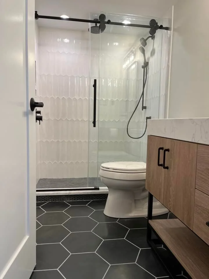 Modern bathroom with a glass shower enclosure, a white toilet, and a wooden vanity with a marble countertop. The floor is covered with large black hexagonal tiles.