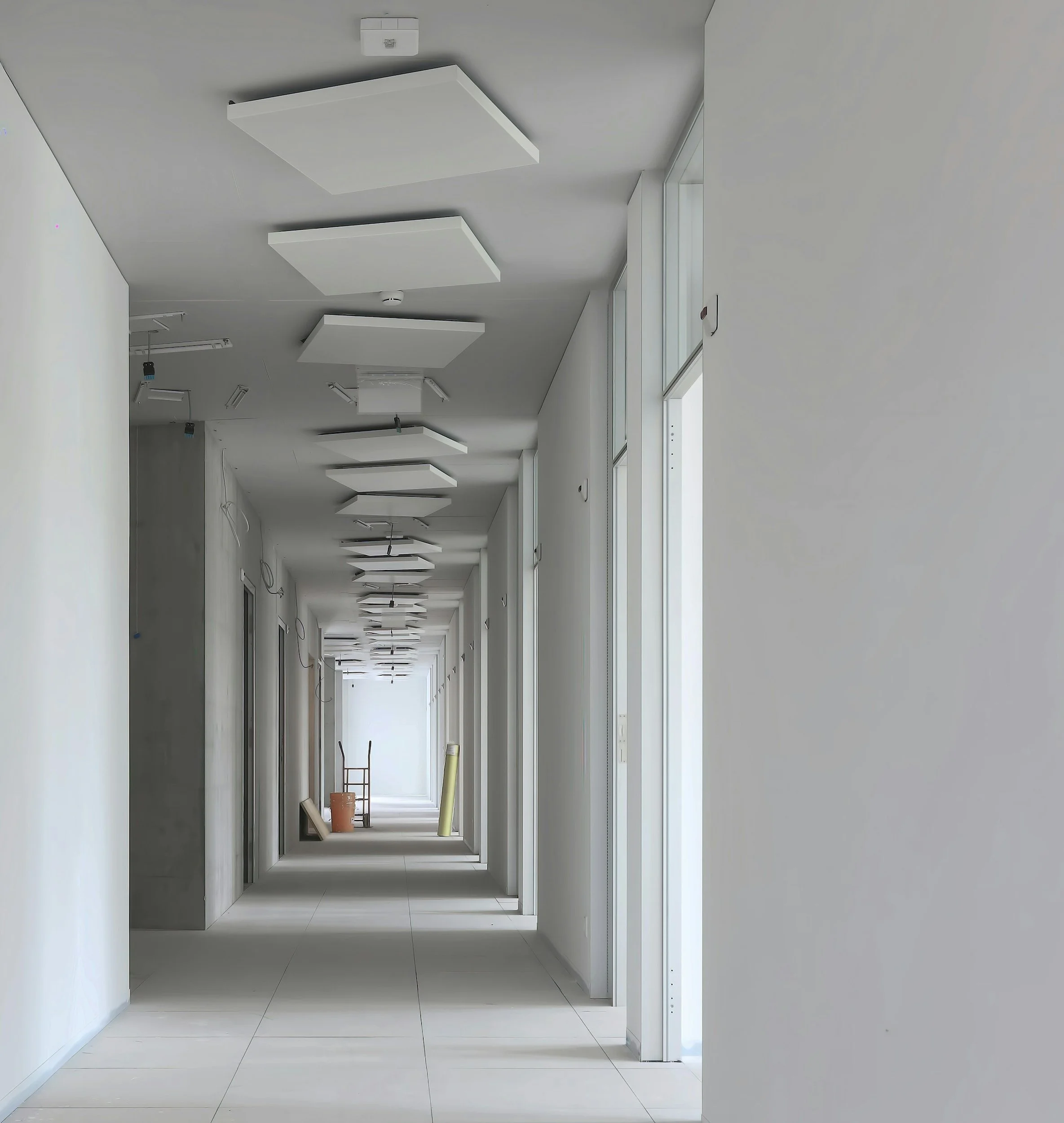Long, white hallway under construction with ceiling panels, construction tools, and materials along the corridor.