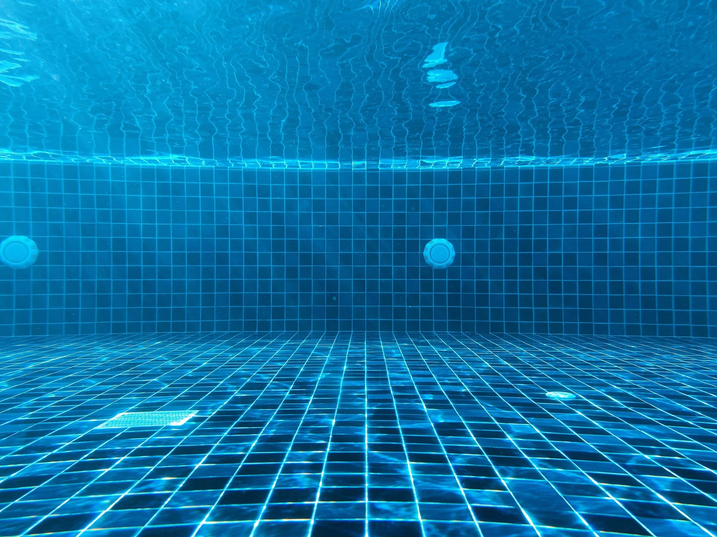 Empty swimming pool with blue tiled walls and floor, underwater view showing pool lights and reflections.