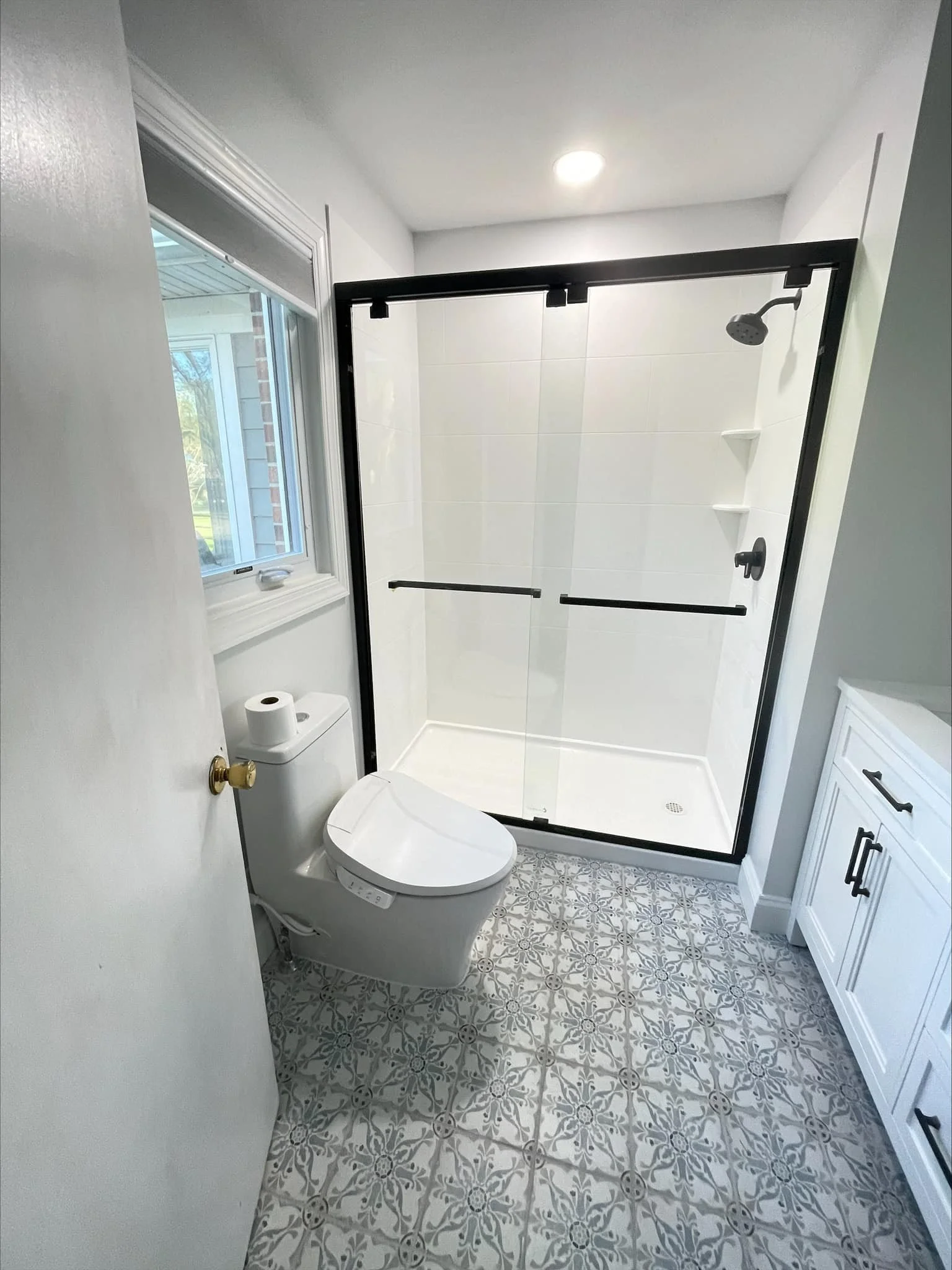 Bathroom with a toilet, a window, a glass shower enclosure with black trim, and a white vanity with black hardware, patterned floor tiles, and a small window on the wall.