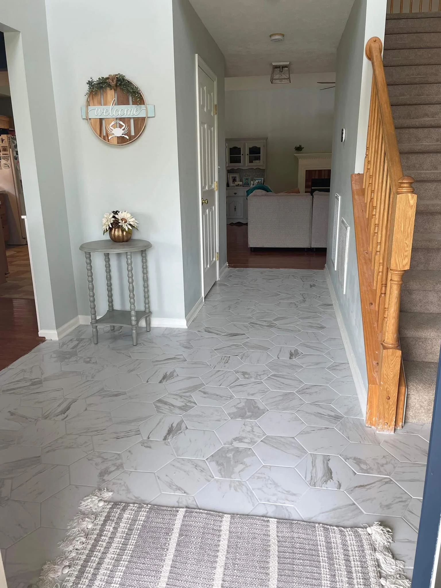 Entryway with white hexagonal marble floor tiles, a small gray side table with a flower vase, and a staircase with wooden railing on the right.