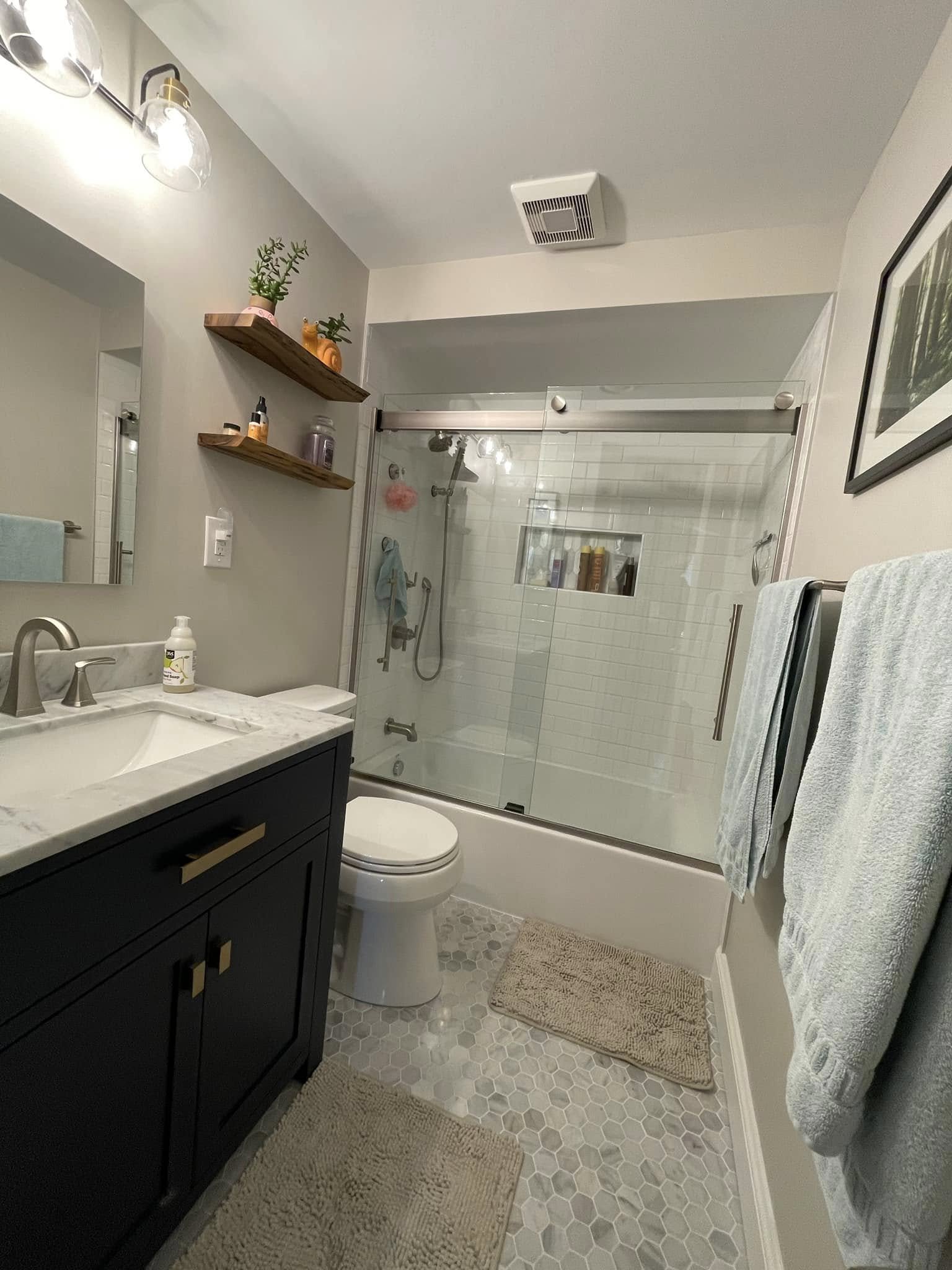 Modern bathroom with a dark vanity, white marble countertop, and glass-enclosed shower with white tile and built-in shelf, beige hexagonal floor tiles, and hanging towels.