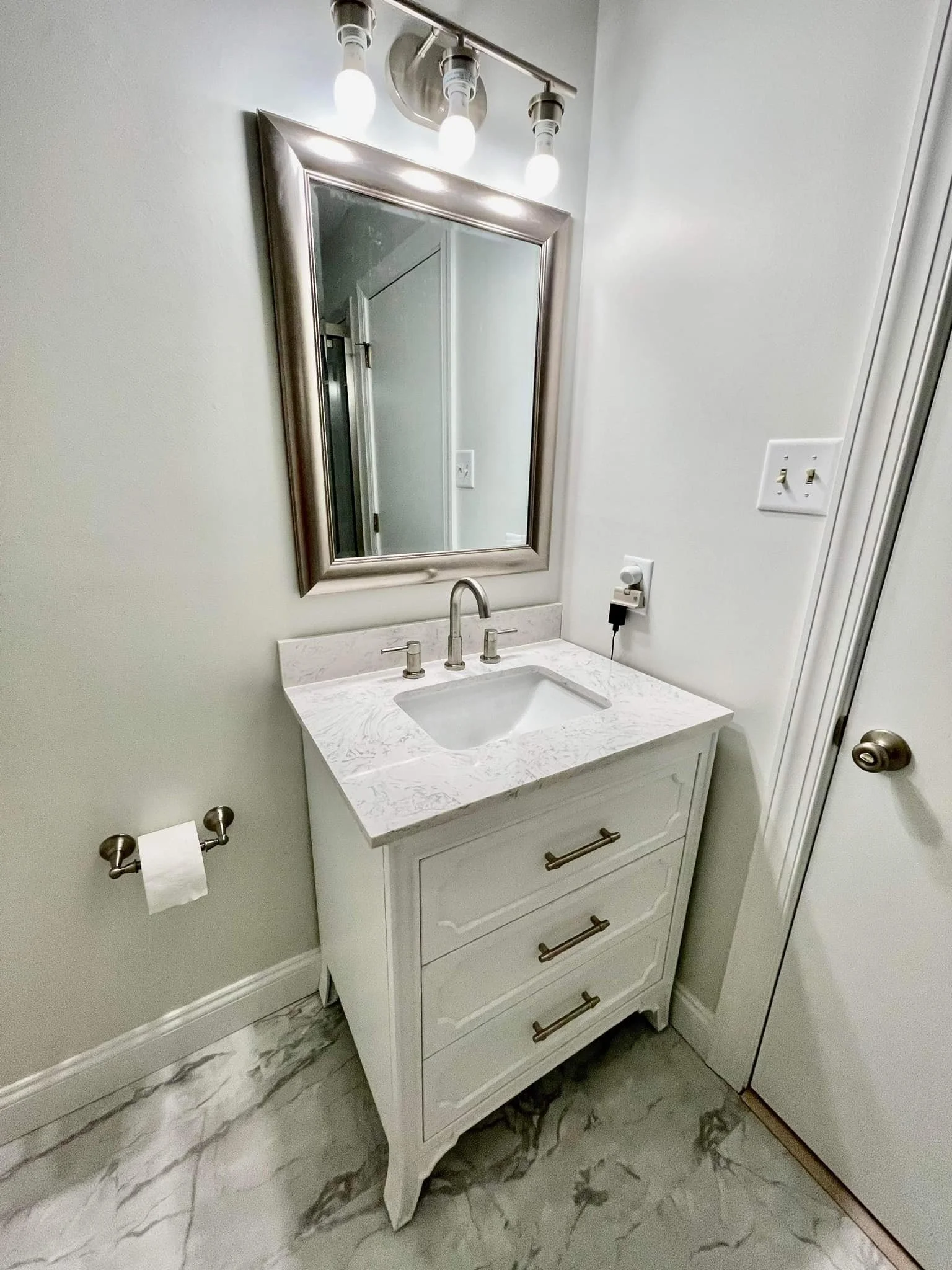 Bathroom vanity with white marble countertop, undermount sink, three drawer cabinet, silver handles, mirror, three light fixtures above, and light-colored tiled floor.