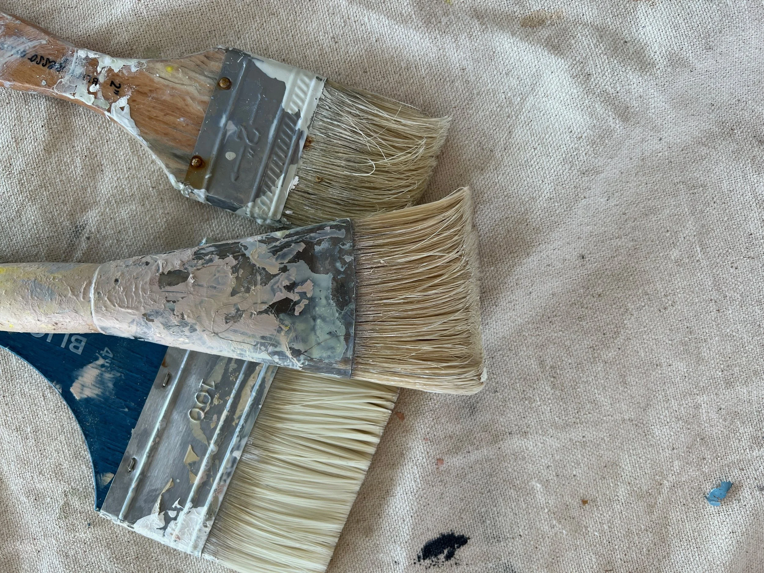 Three paintbrushes with dried paint on their handles and bristles lying on a beige canvas surface.