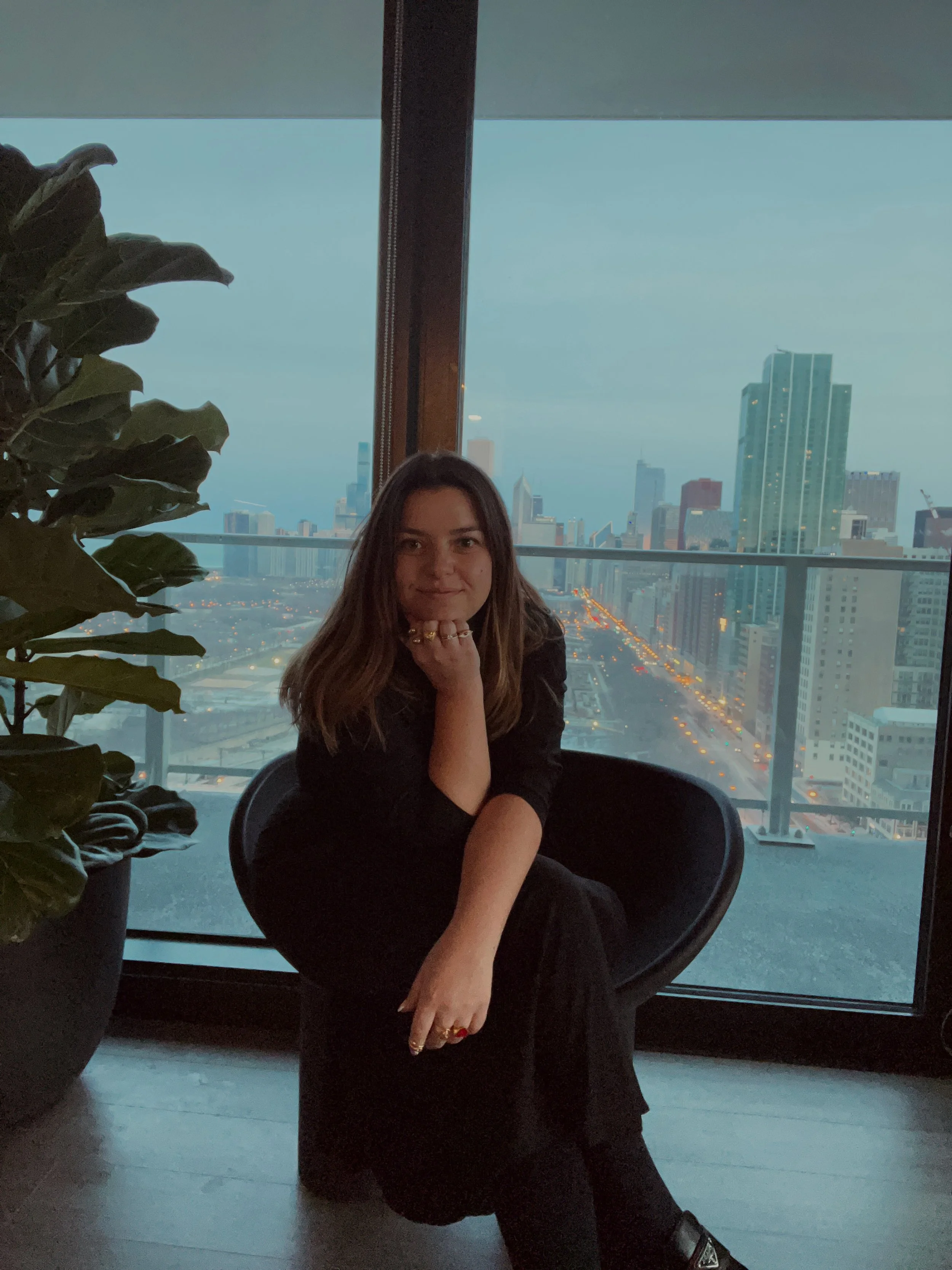 Young woman with brown hair sitting in a black chair, posing with her chin resting on her hand, in front of a large window showing a city skyline at dusk.