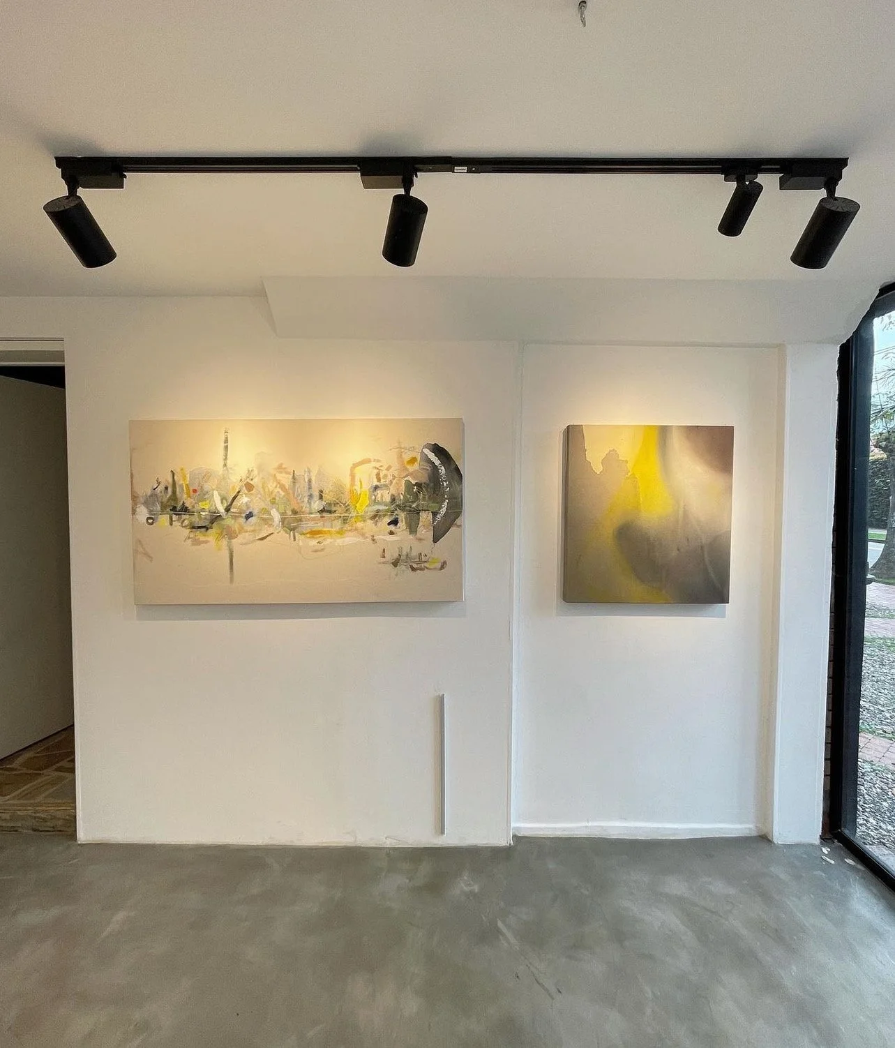 An art gallery showing two abstract paintings on a white wall, with track lighting on the ceiling and a glass door on the right.