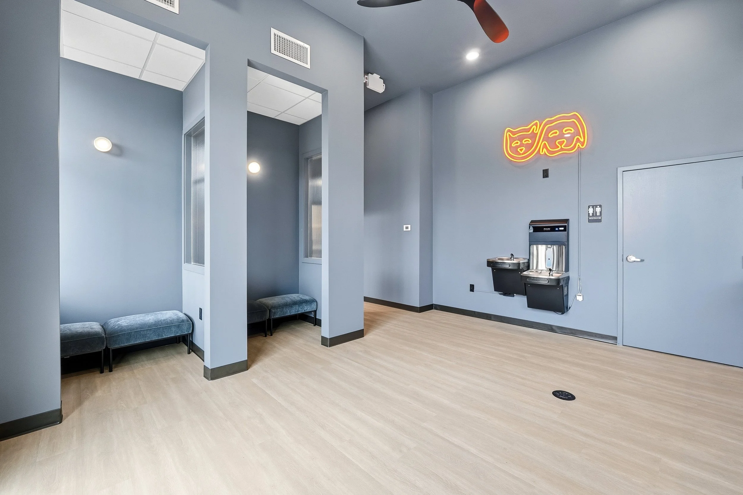 Modern waiting area facing inside with two small semi-private waiting booths containing padded benches, wall-mounted water fountains, orange neon logo sign, and a bathroom. 