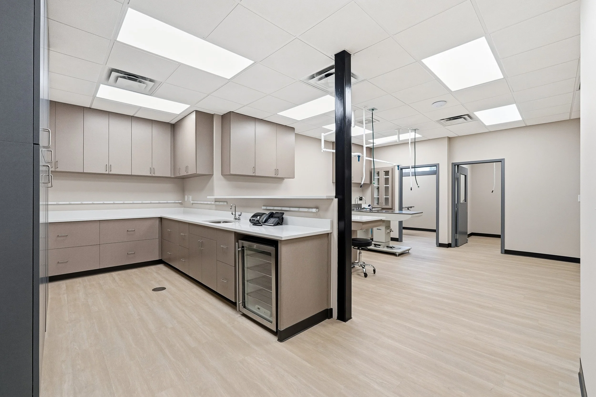 Medical laboratory with beige cabinets, a sink, a phone, a mini fridge, and medical equipment, with bright fluorescent lighting and light-colored wood flooring.