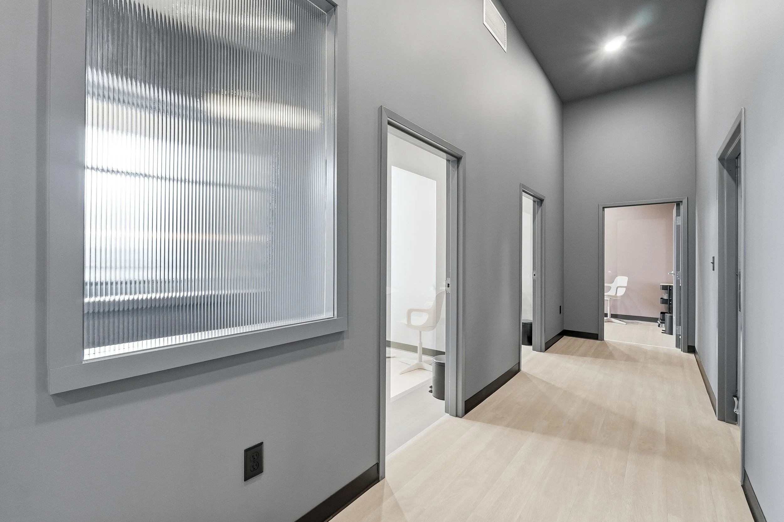 An open hallway with multiple doorways leading to private exam rooms, light wood flooring, and a large textured glass interior window next to one of the waiting cubbies.