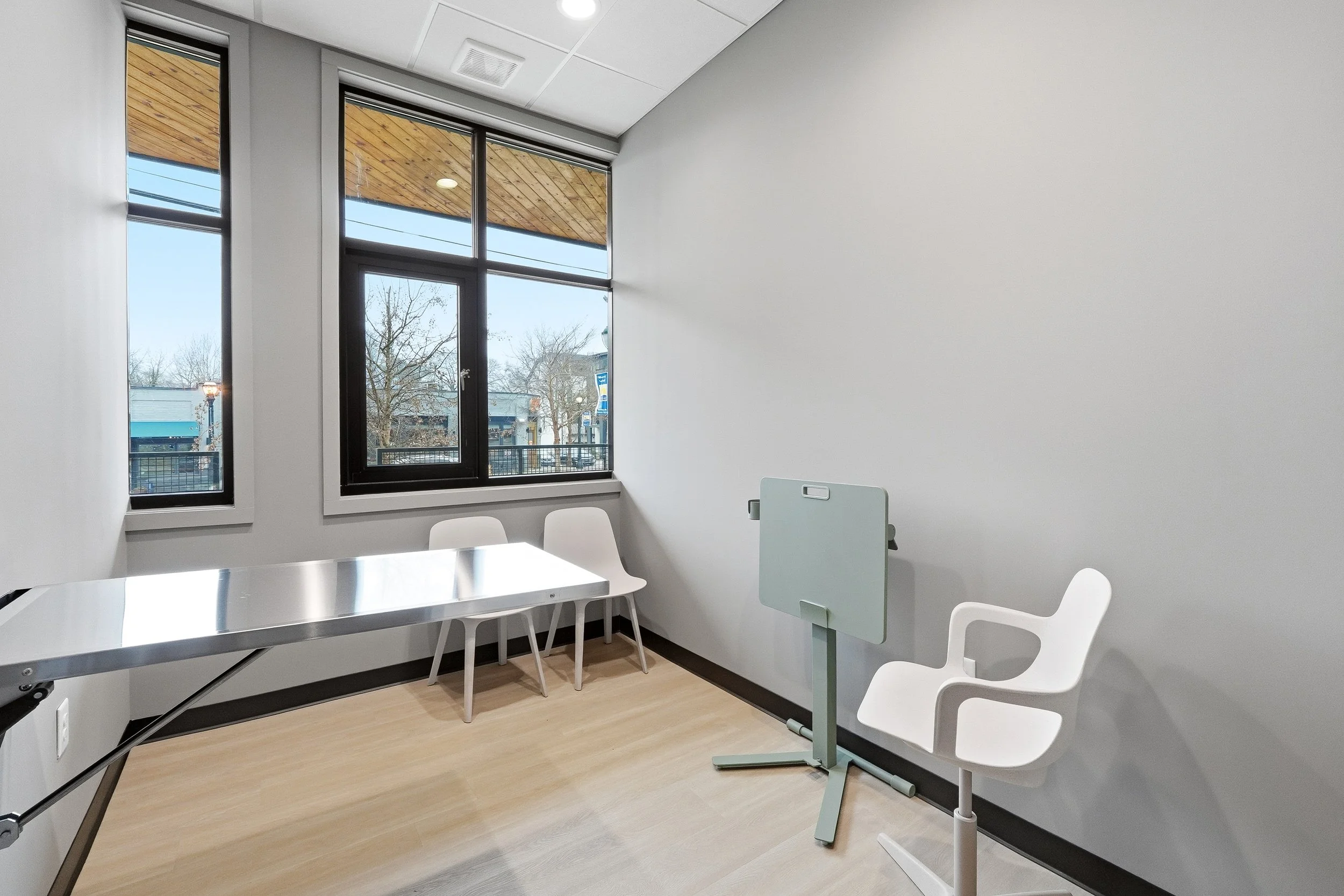 Exam room with large windows, two chairs along the windows, a chair and mobile desk for a team member, and a folding exam table.