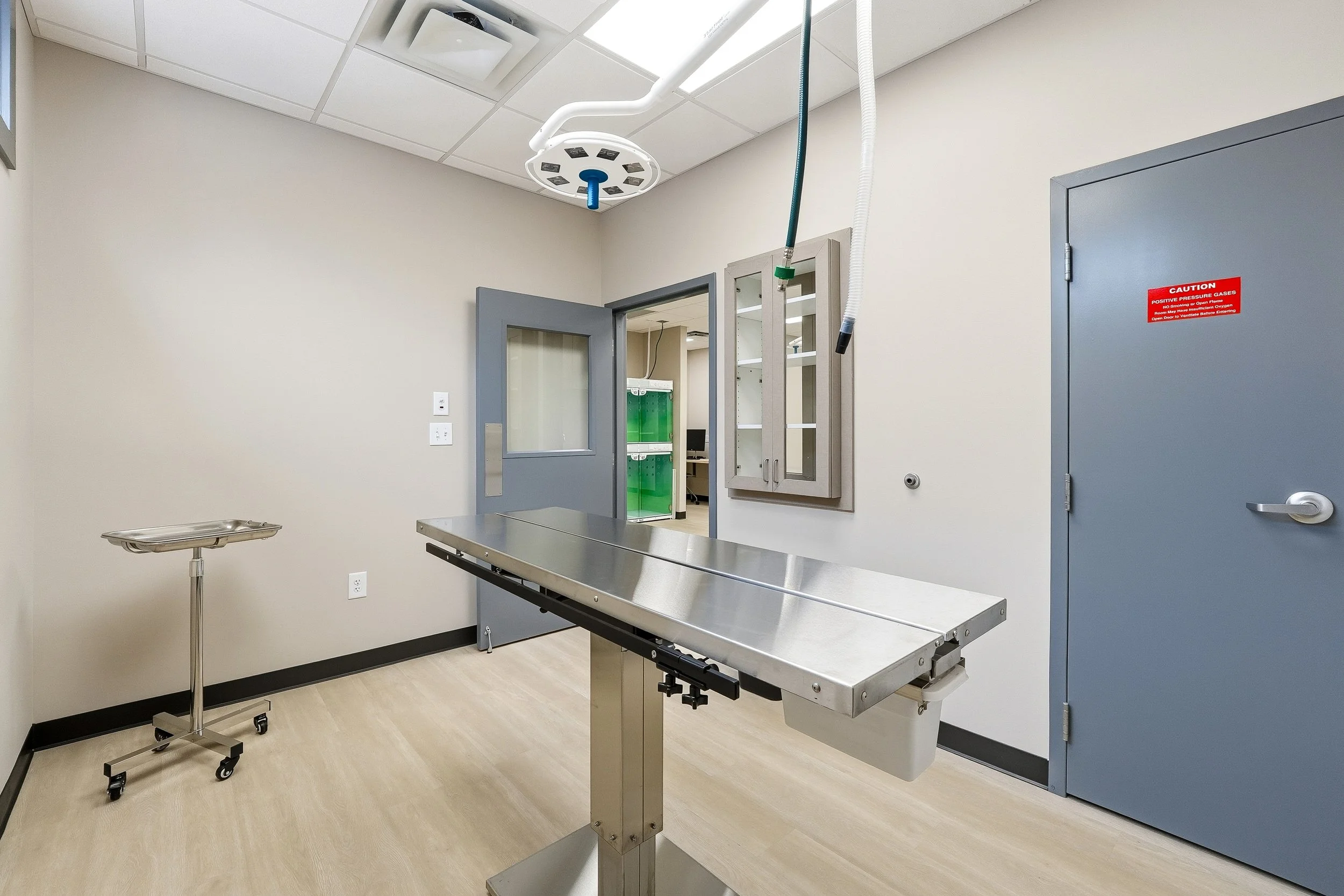 A sterile medical surgery room with beige walls, a stainless steel, heated, lifting examination table, a small rolling tray table, an overhead surgical light, and a pass-through cabinet with glass doors. Not pictured is the anesthesia machine. 