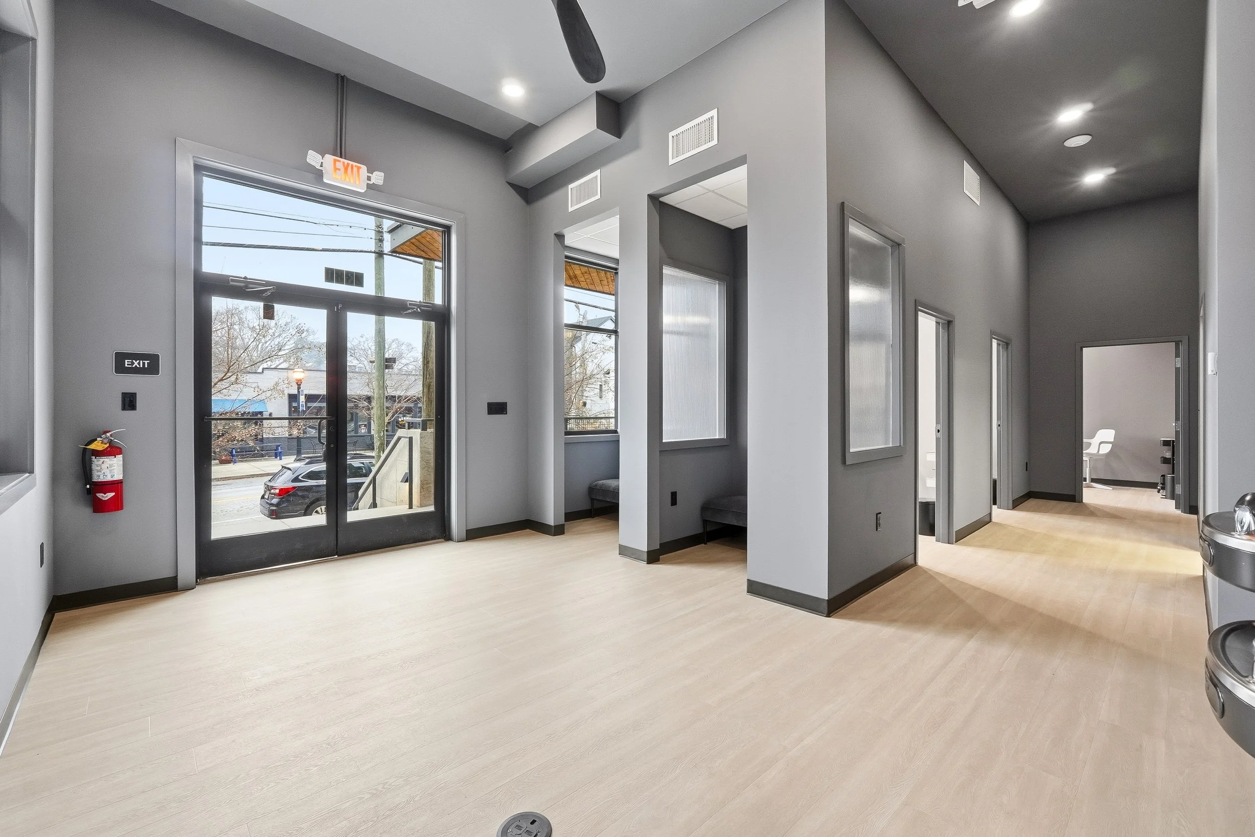 Waiting area from the perspective of the bathroom door; toward the left is looking at the waiting area; light wood floors, large glass entrance door, and multiple rooms down the hallway to the right