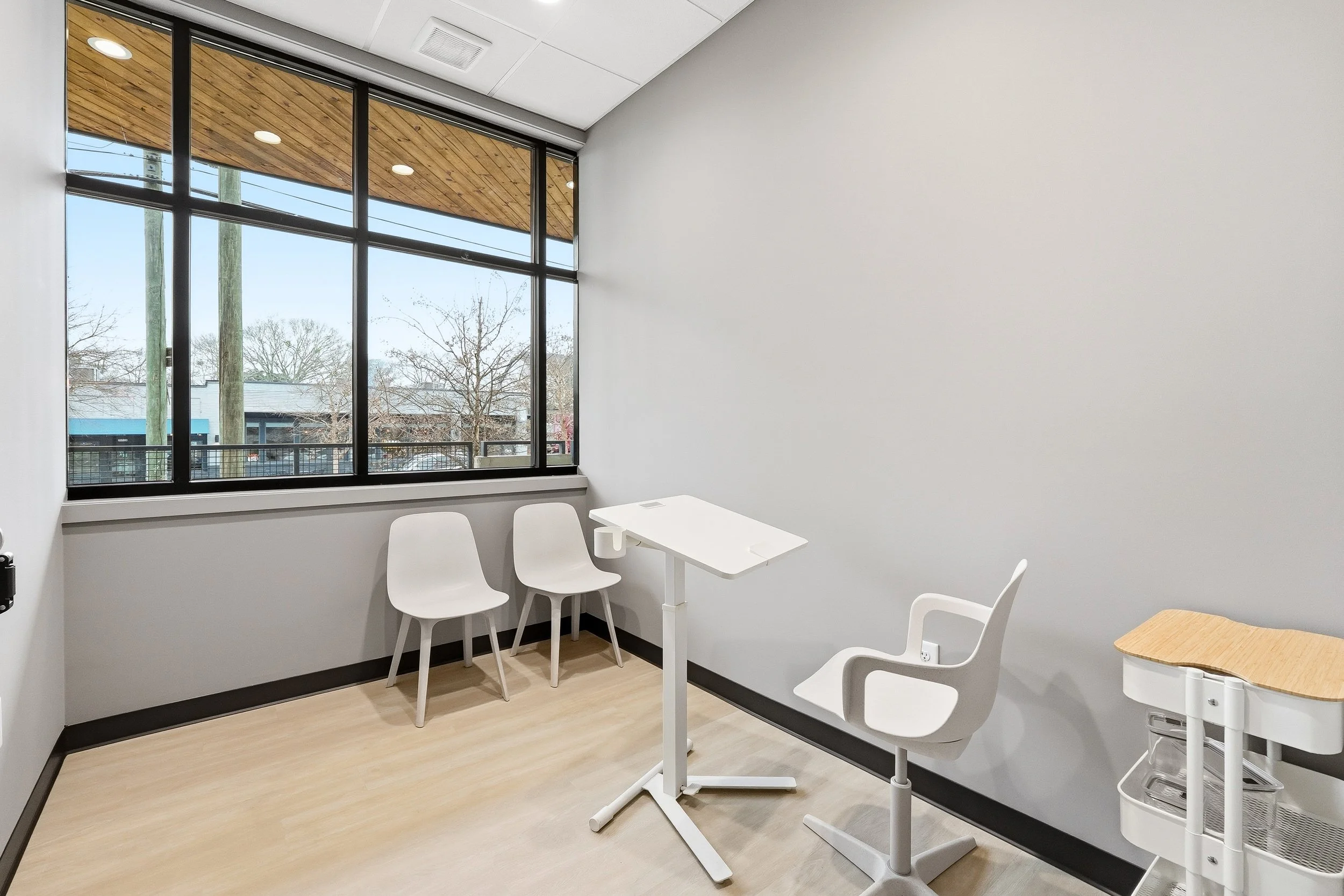 Exam room with large windows, two chairs along the windows, a chair and mobile desk for a team member, and a folding exam table.