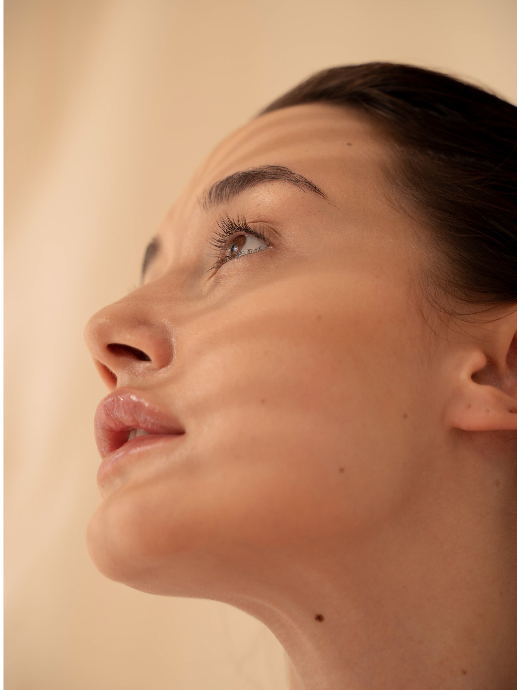 Close-up of a woman's face from a low angle, showing her upward gaze, smooth skin, and dark hair pulled back.