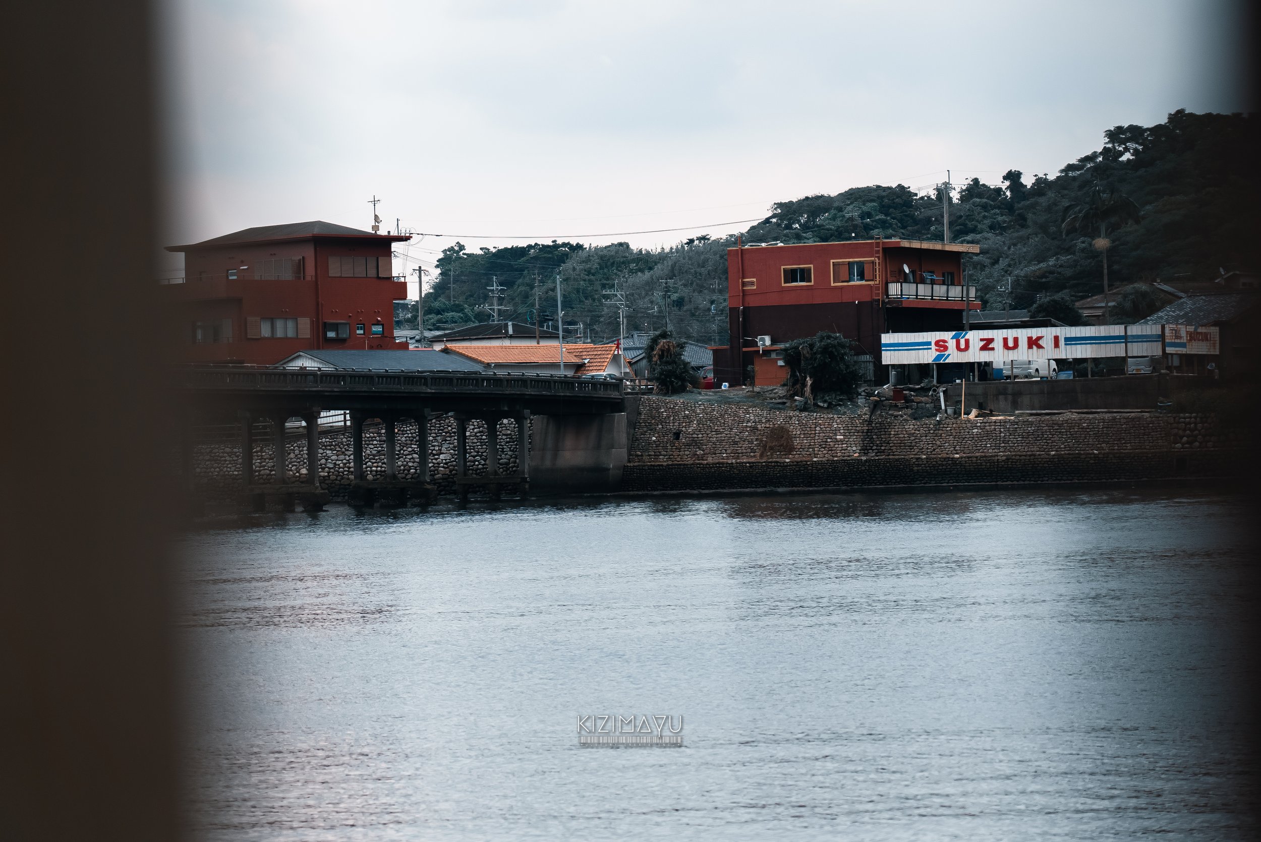 川辺に建つ赤い建物と橋、遠くに山と空。