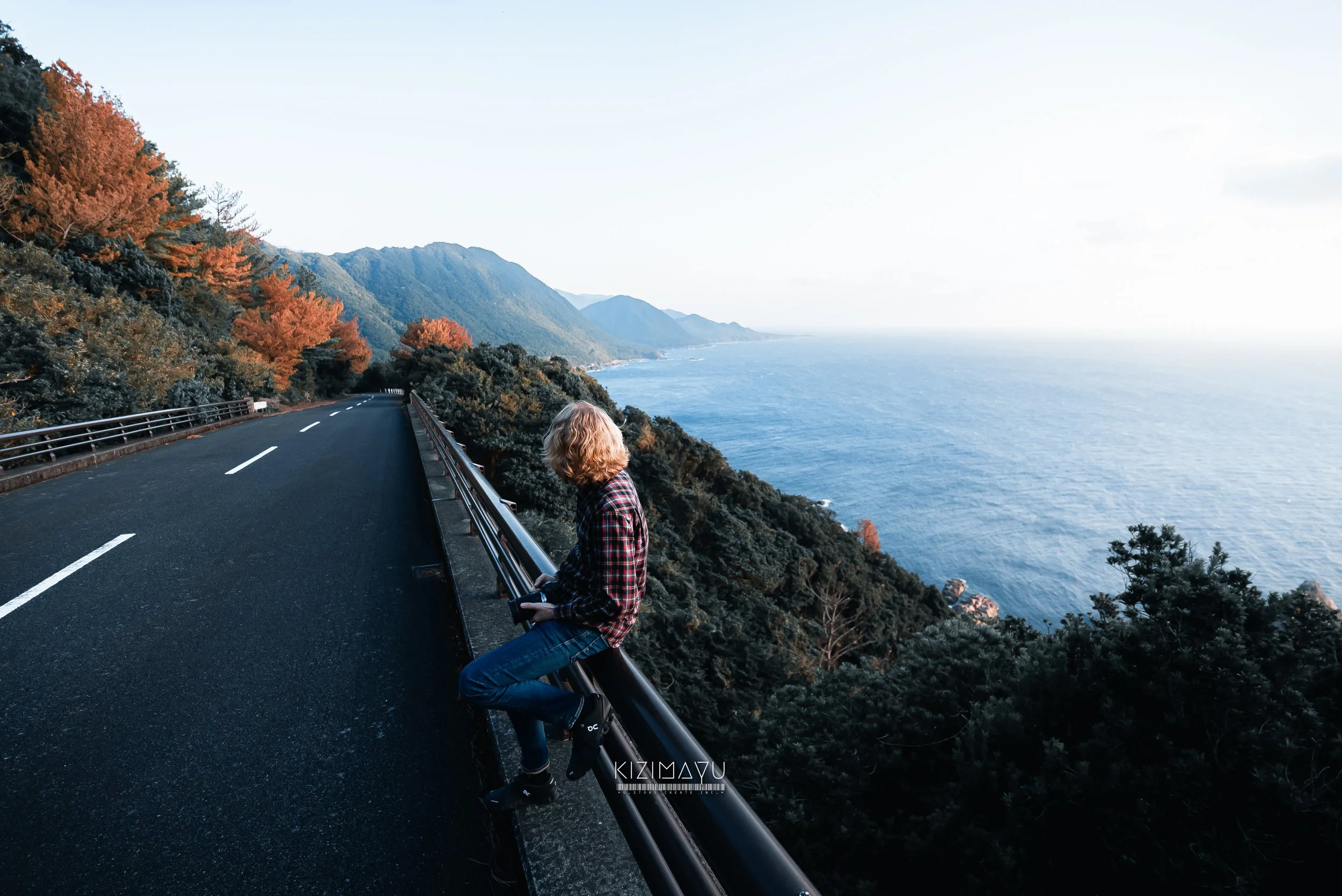 海に面した山道にいる女性。左側に道路と道路脇のガードレール。背景に青い海と山々。女性はチェック柄シャツとジーンズを着て、座っている。秋の色づいた木々が左右に見える。