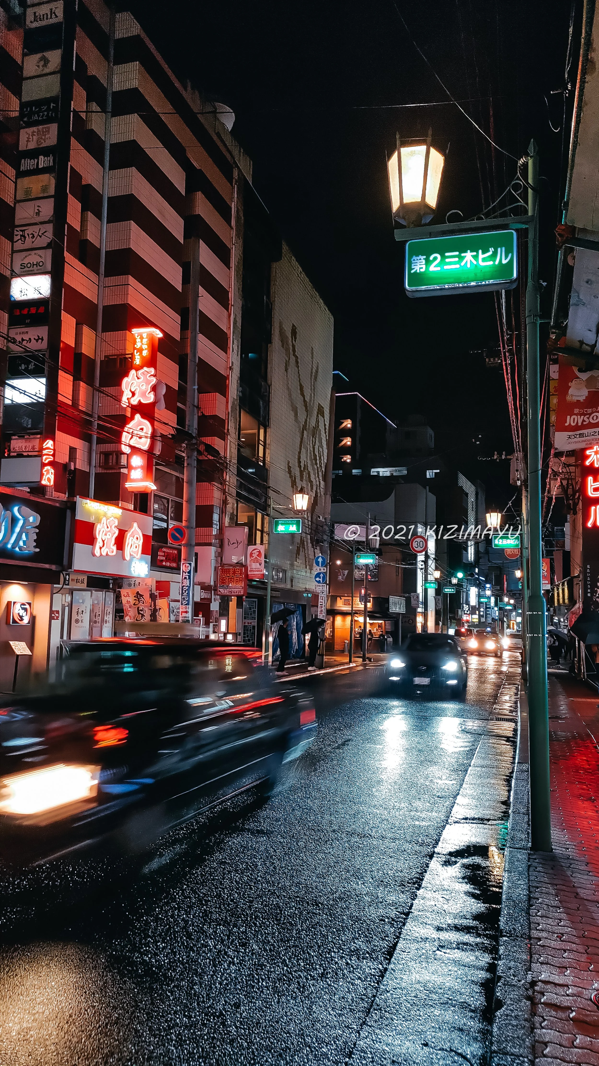 夜の雨にぬれた街の通り、車のライトとネオンサインが輝く。左側にレストランの看板、右側に第2三木ビルの看板が見える。