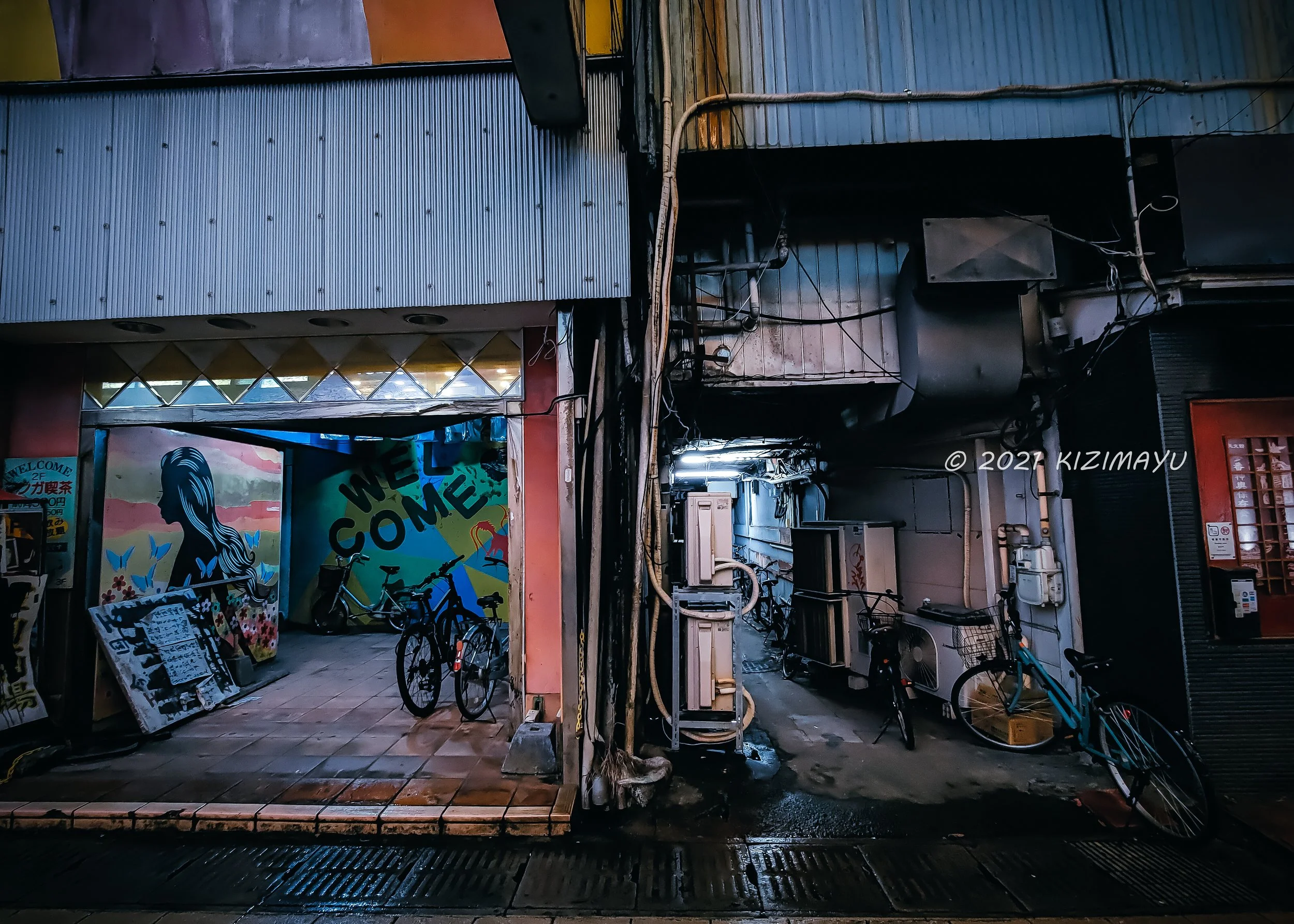夜の街角にあるバイクと自転車、壁に描かれた女性のイラストとメッセージが書かれた壁画が見える。