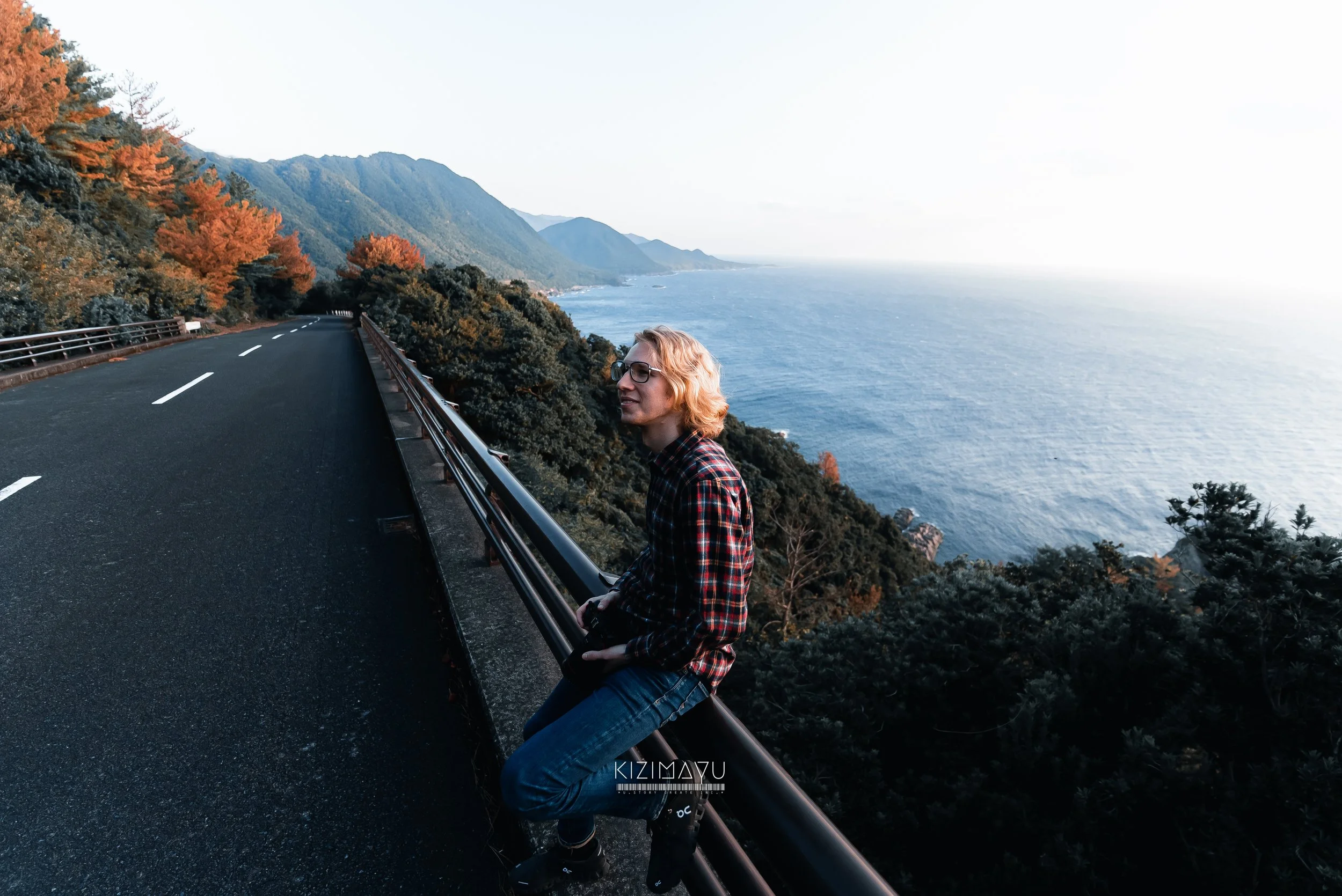 海沿いの山道を走る車の側で座る若い男性、右側に海と山々の景色が広がる風景