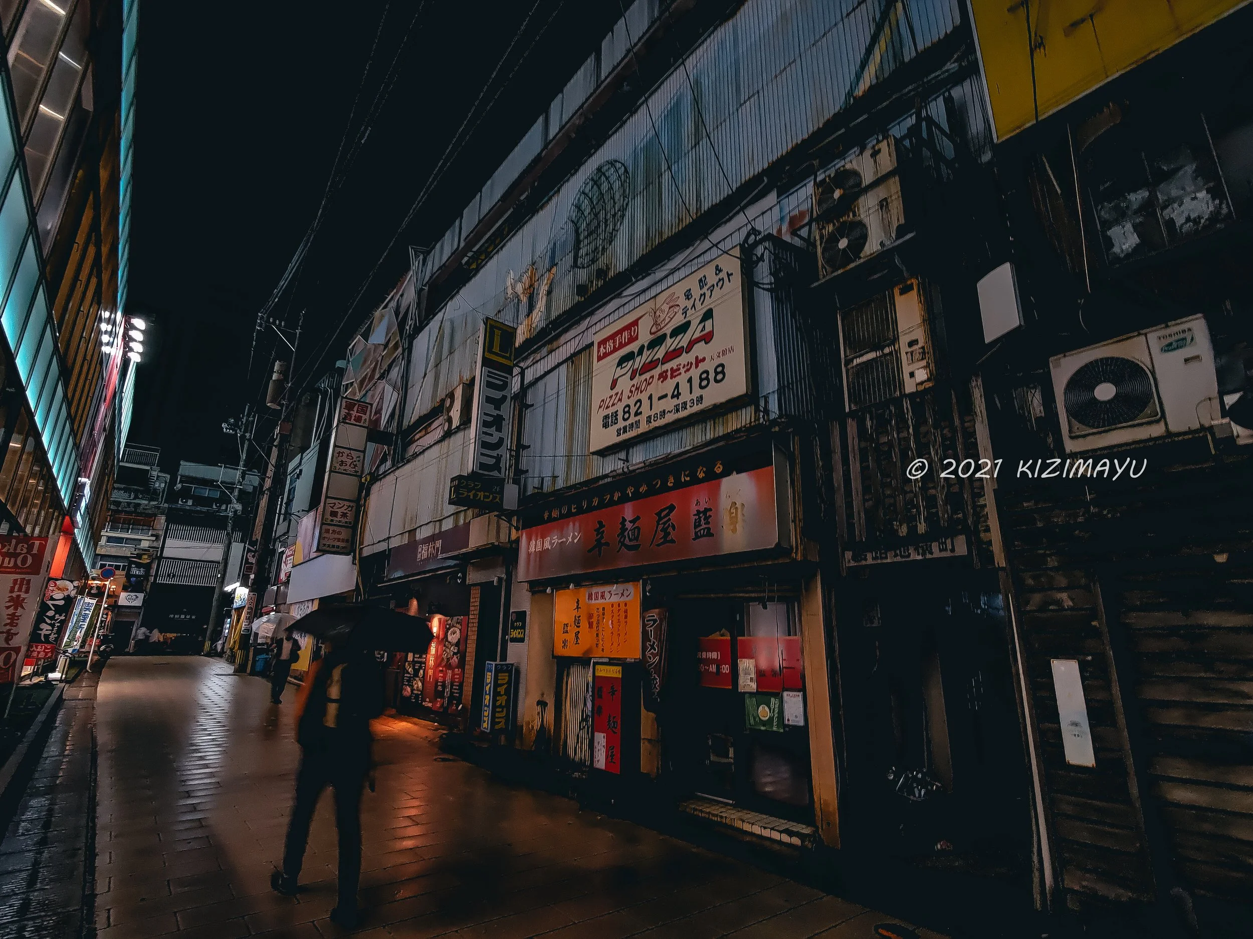 夜の街の通りにある飲食店や看板、傘をさした人々。舗装された通路と営業中の店舗。